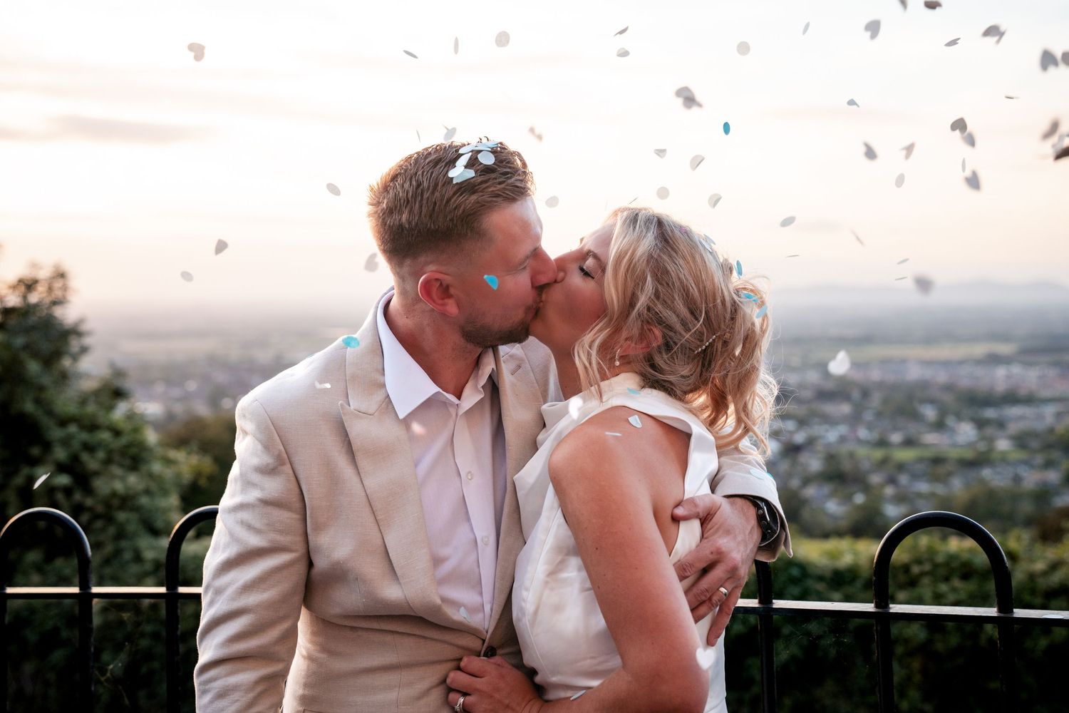 Newlywed couple sharing a romantic kiss outdoors with confetti falling, groom in beige suit, bride in white dress.