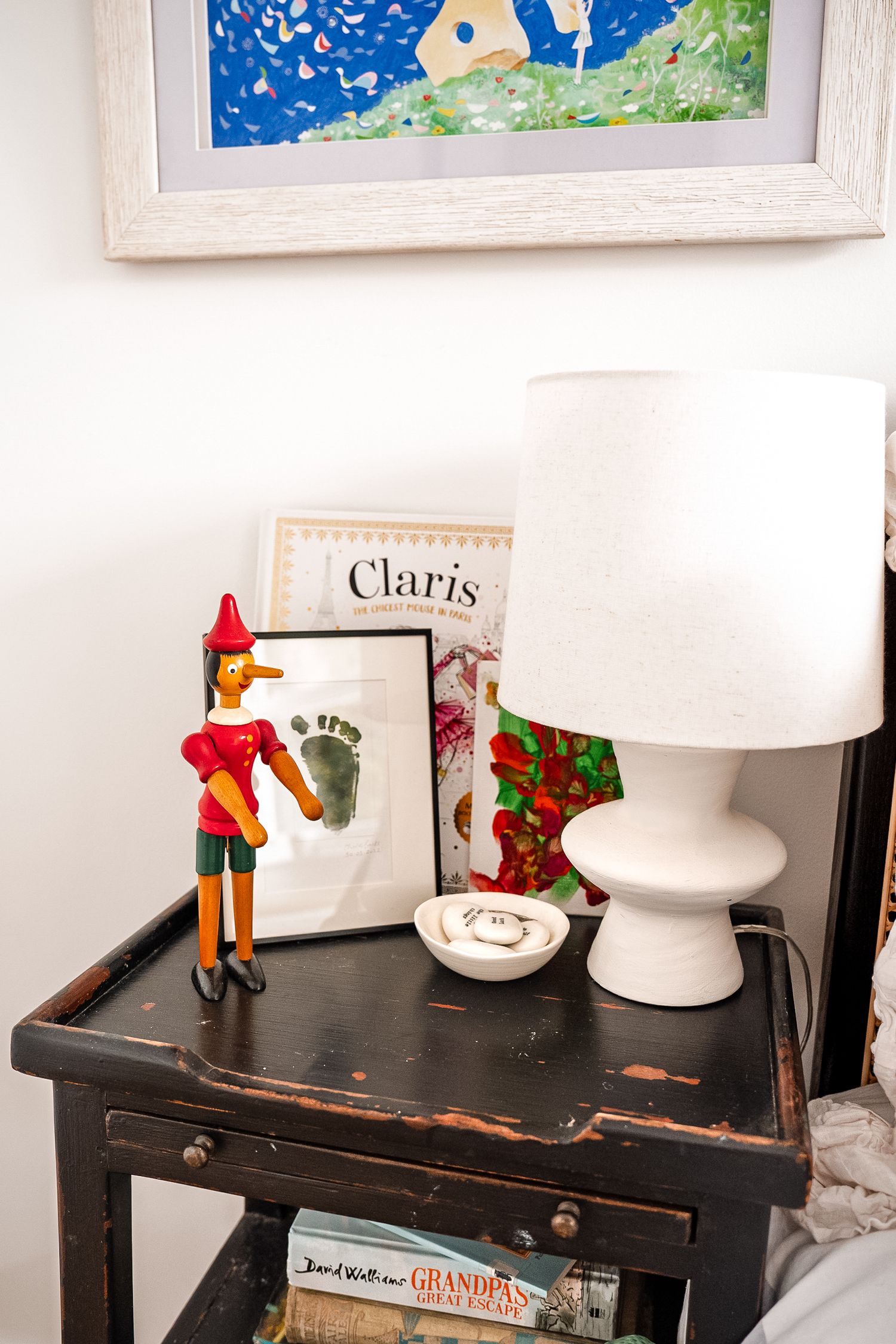 Vintage black nightstand with Pinocchio figurine, white ceramic lamp, Claris book, and small trinket dish.