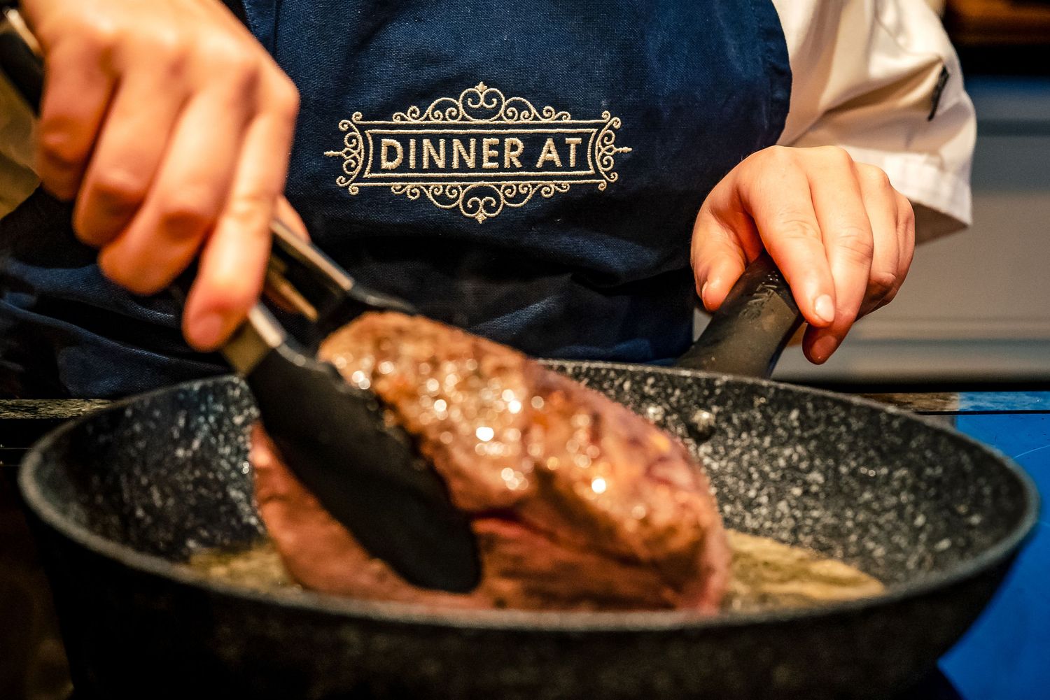 Chef in a Dinner At apron searing a steak in a dark non-stick pan with tongs.