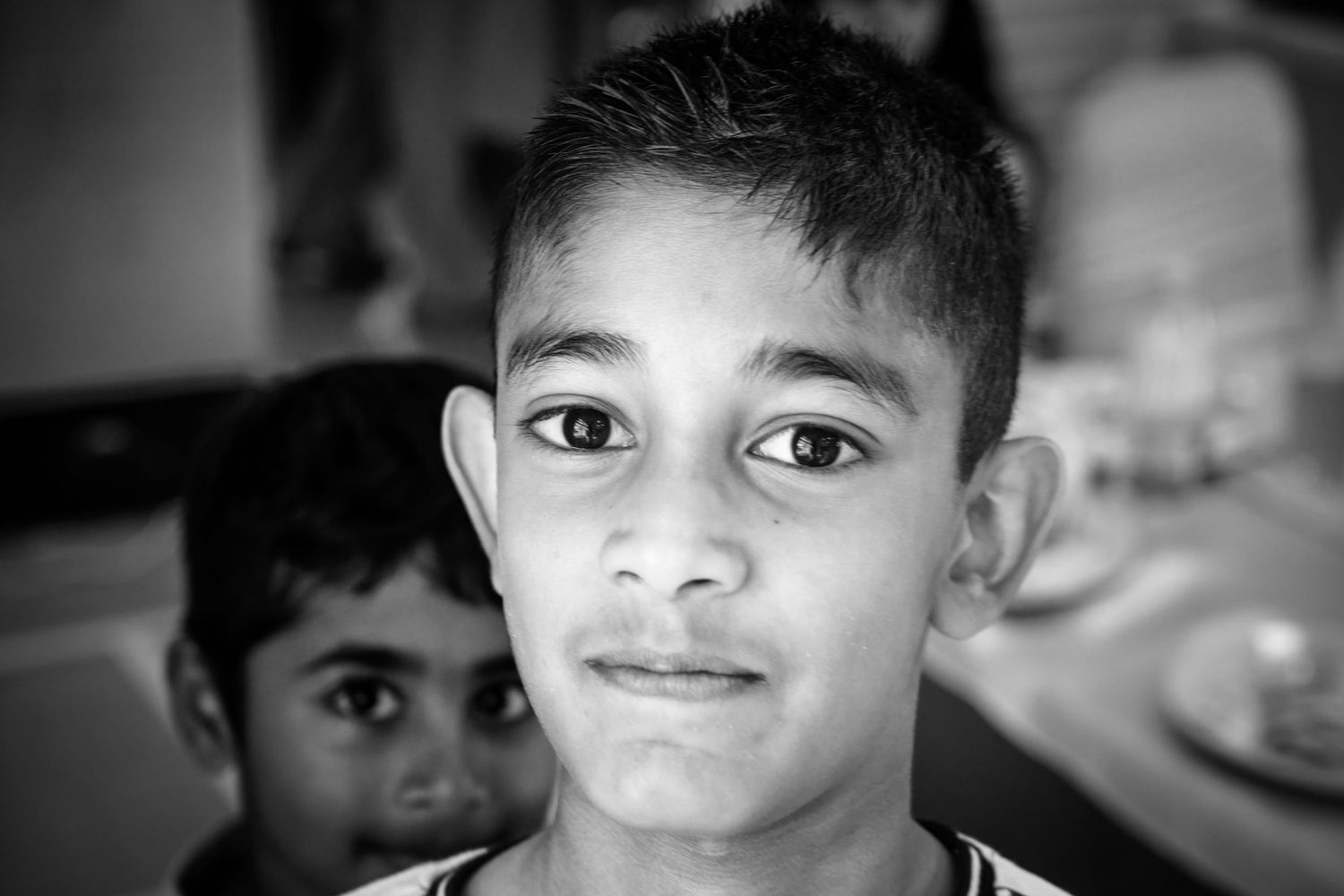 Black and white portrait of a young boy looking at camera with another child visible in the background.