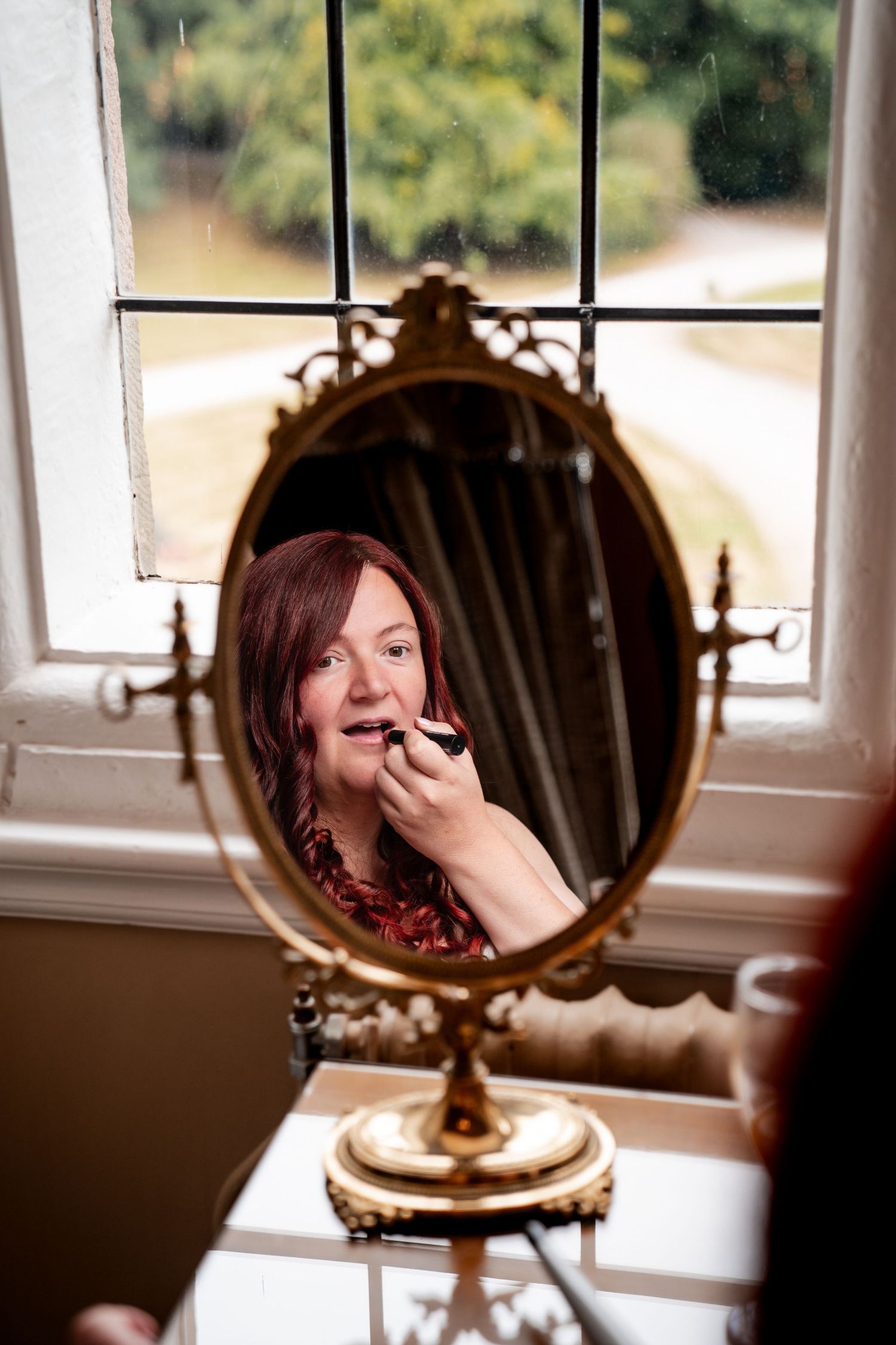 Woman applying lipstick reflected in an ornate gold oval mirror near a window.