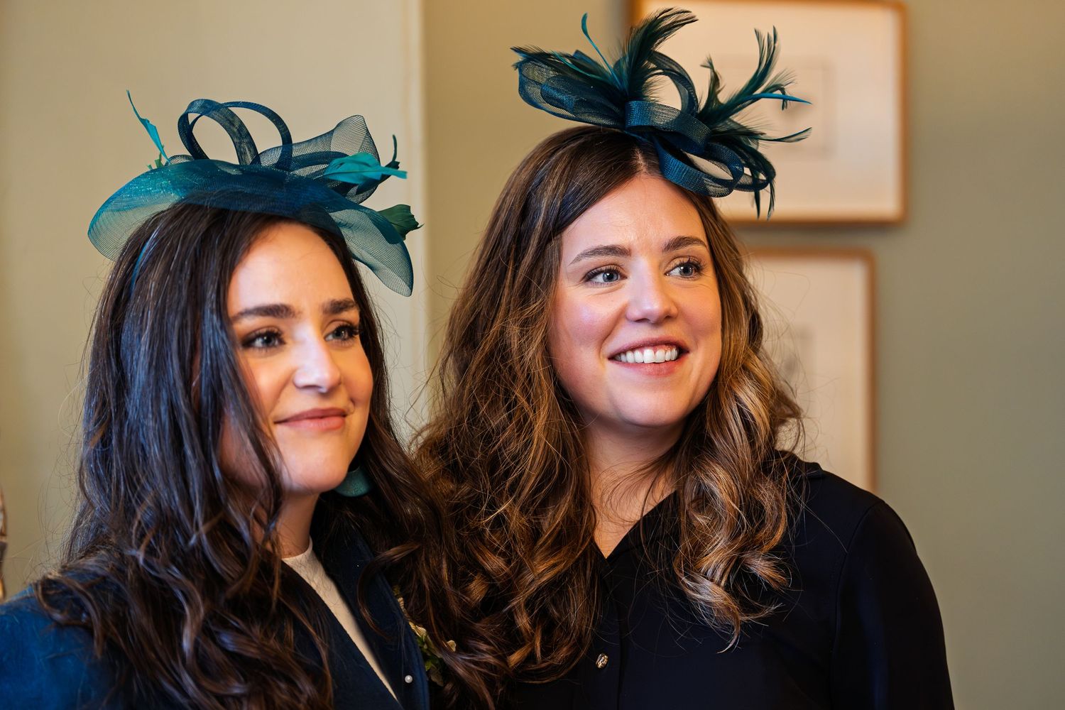 Two women wearing matching teal fascinator hats with feathers, smiling indoors at a formal event.