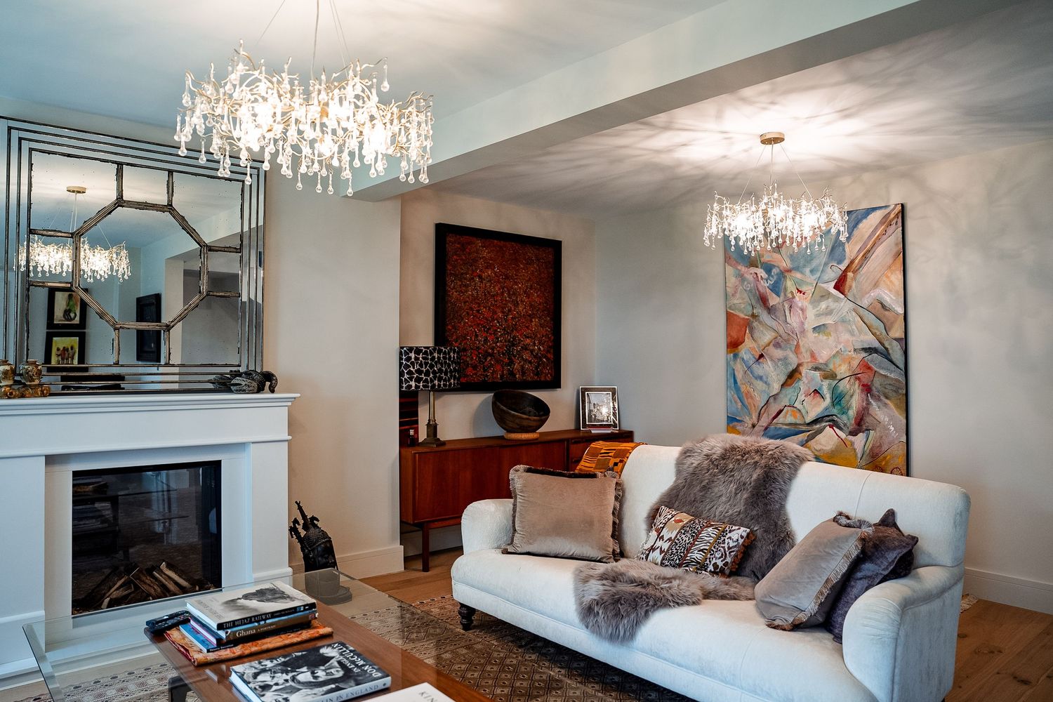 Elegant living room with white sofa, crystal chandeliers, fireplace, abstract artwork, and fur throw pillows on hardwood floors.