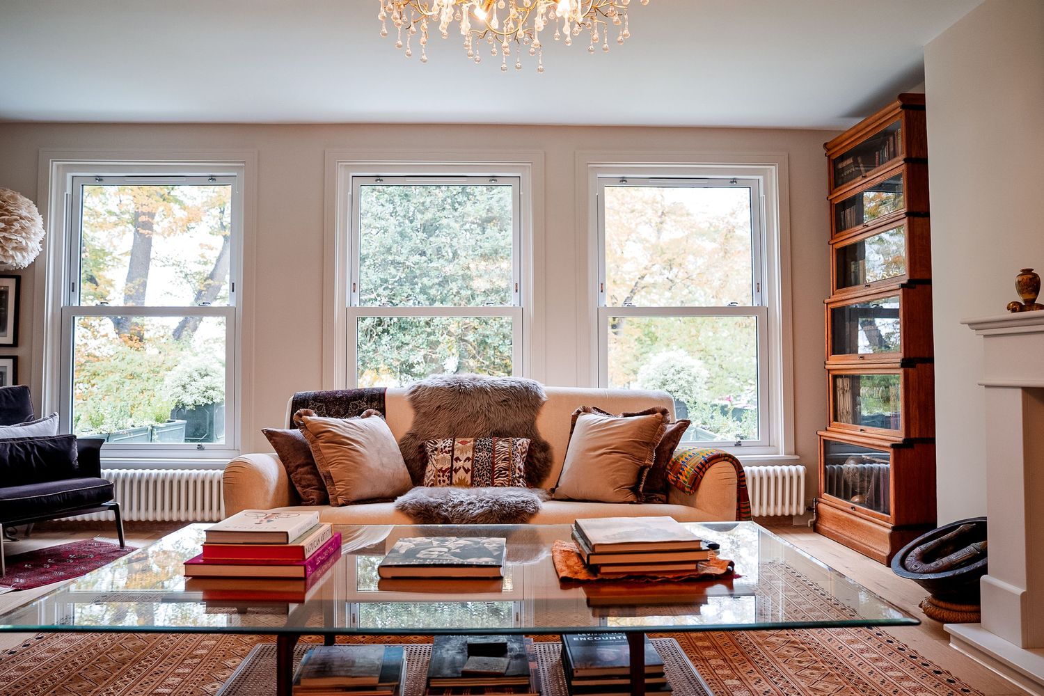 Elegant living room with tan sofa, glass coffee table, bookshelf, and large windows letting in natural light.