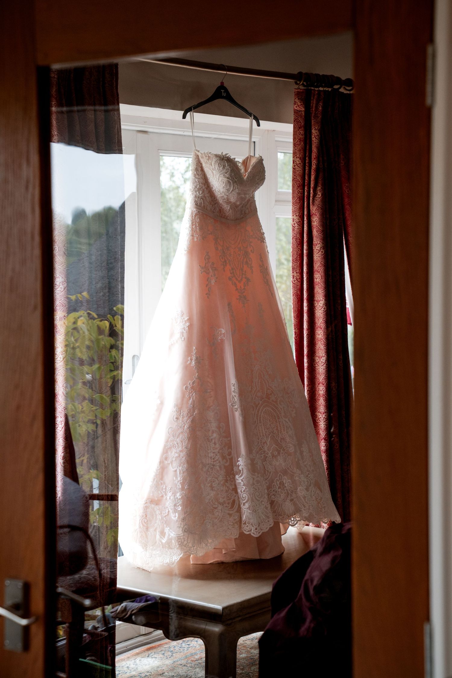 A elegant blush lace wedding dress hanging on a hanger in front of a sunlit window with dark red curtains.