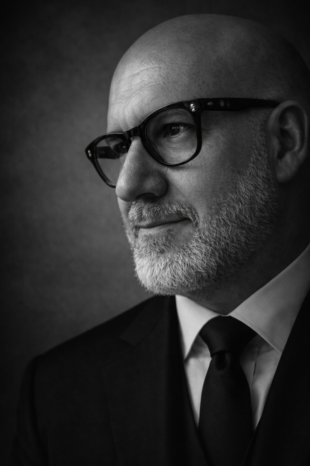 Black and white portrait of a bald man with beard wearing glasses and a suit with tie.