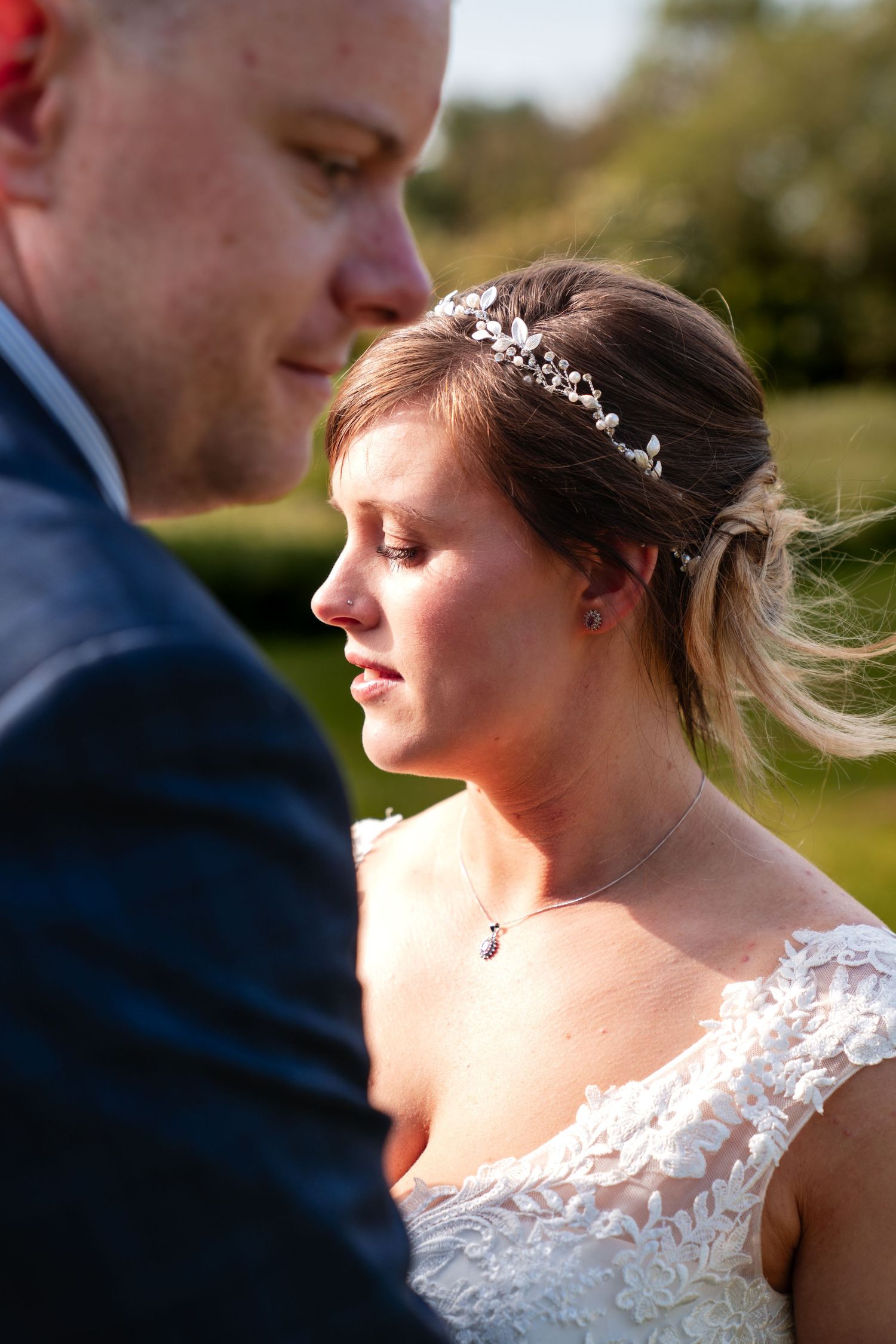 Bride in lace gown and floral hair piece shares an emotional moment with groom in blue suit outdoors.