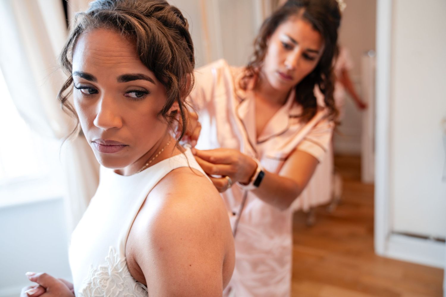 Bridesmaid helping bride put on necklace while getting ready for wedding ceremony.