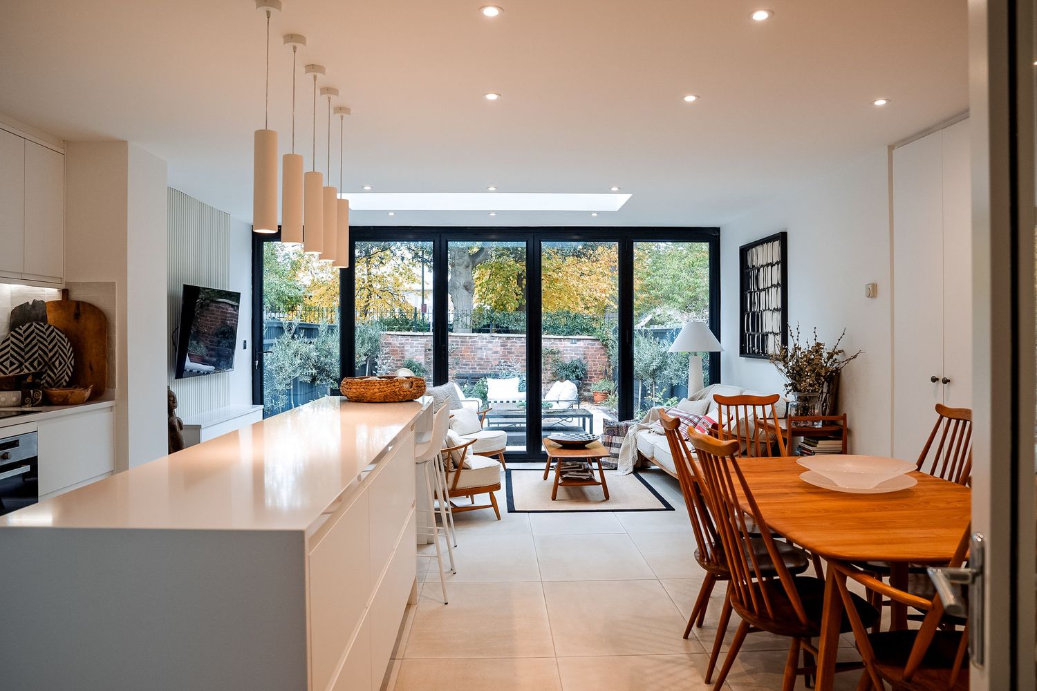 Modern open-plan kitchen, dining, and living area with white cabinetry, wooden dining table, and large glass doors to garden.