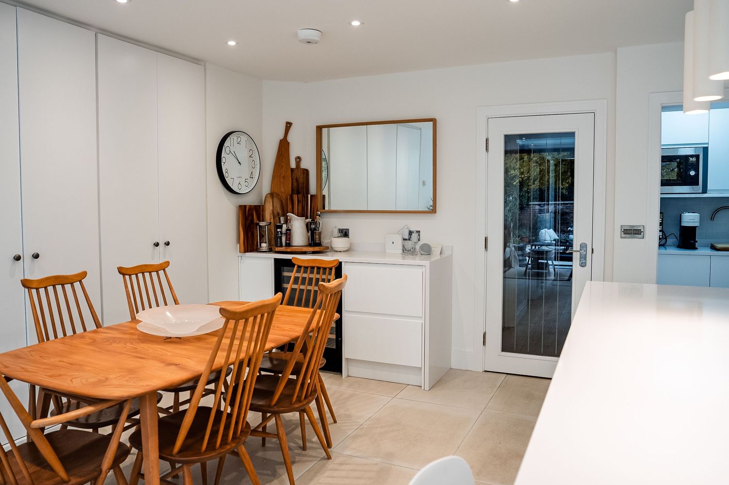 Modern white kitchen-diner with wooden dining table, chairs, wall clock, and glass door leading to garden.