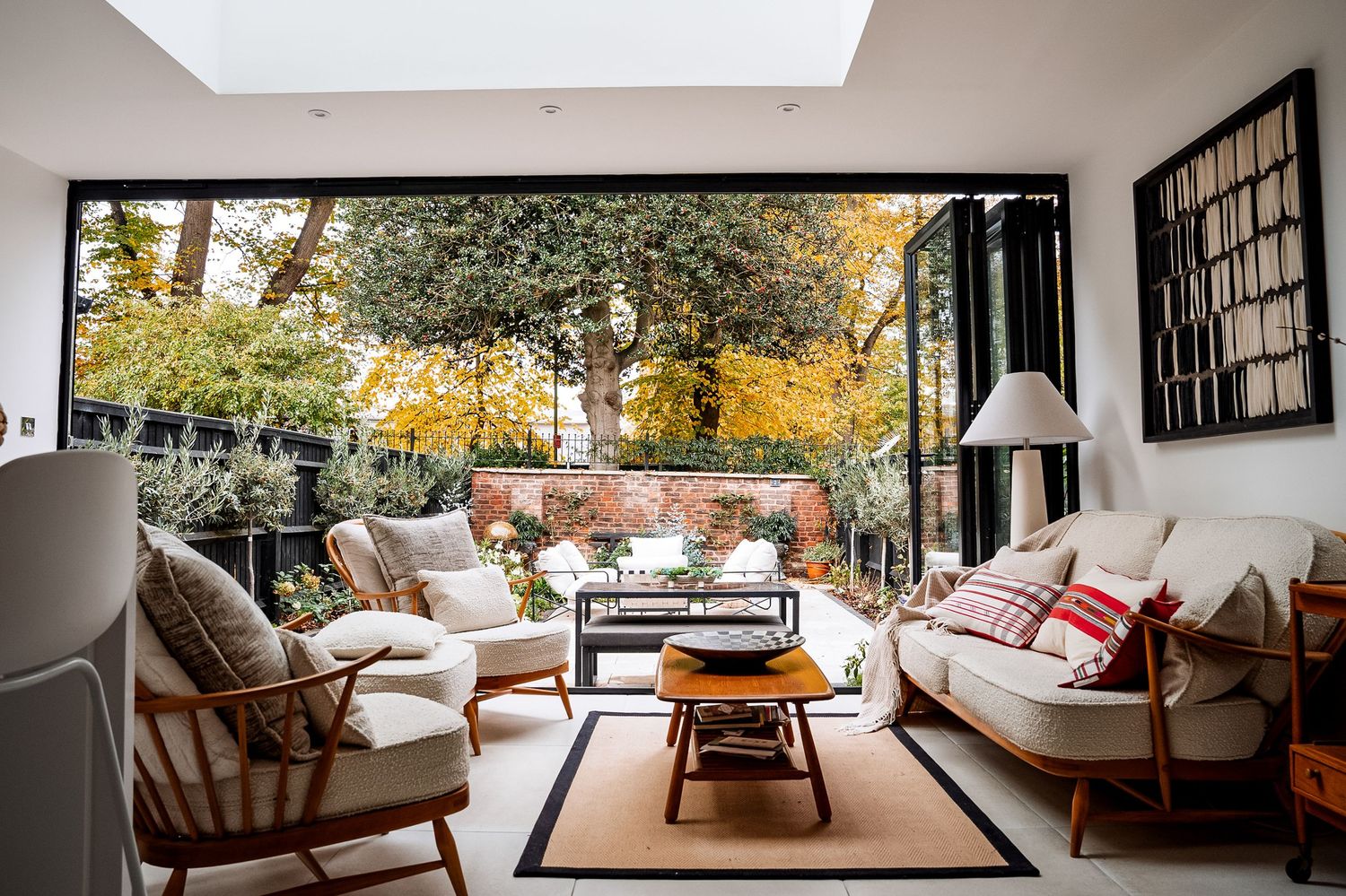 Modern living room with open bifold doors leading to an autumn garden, featuring Ercol furniture and a skylight.