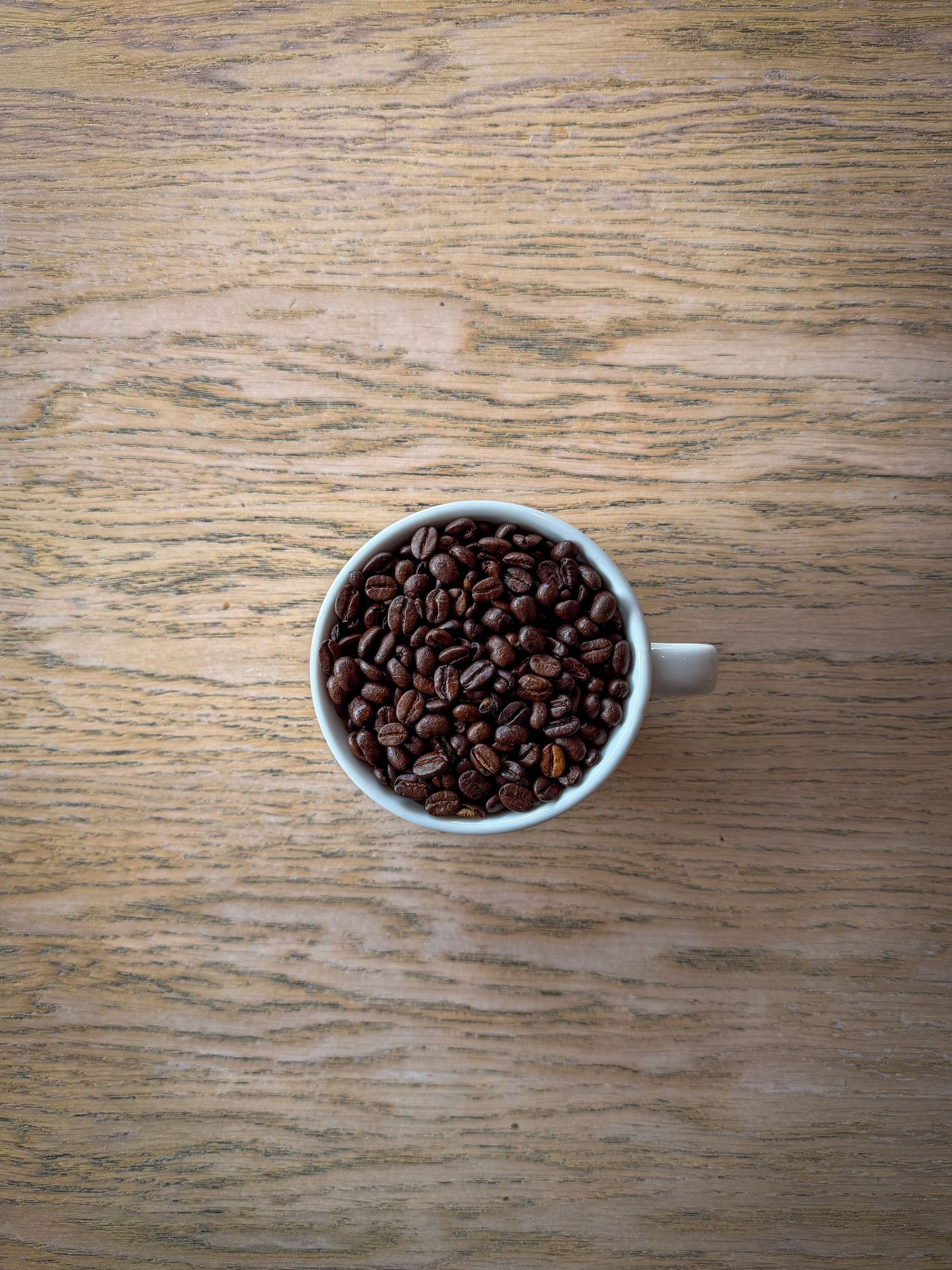 A white ceramic cup filled with roasted coffee beans placed on a wooden table, viewed from above.