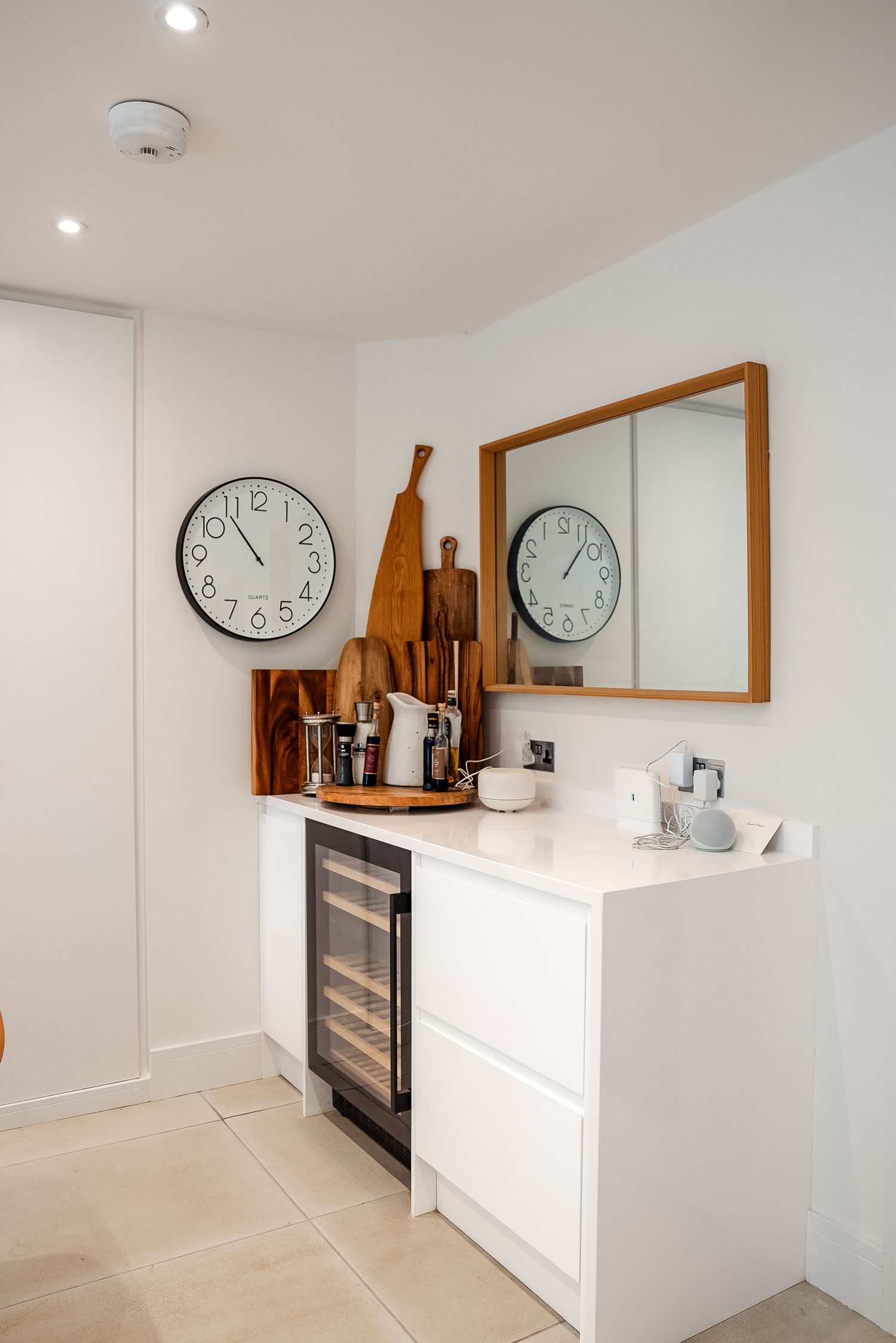 Modern white kitchen counter with wine fridge, wall clock, wooden cutting boards, and framed mirror in bright interior.