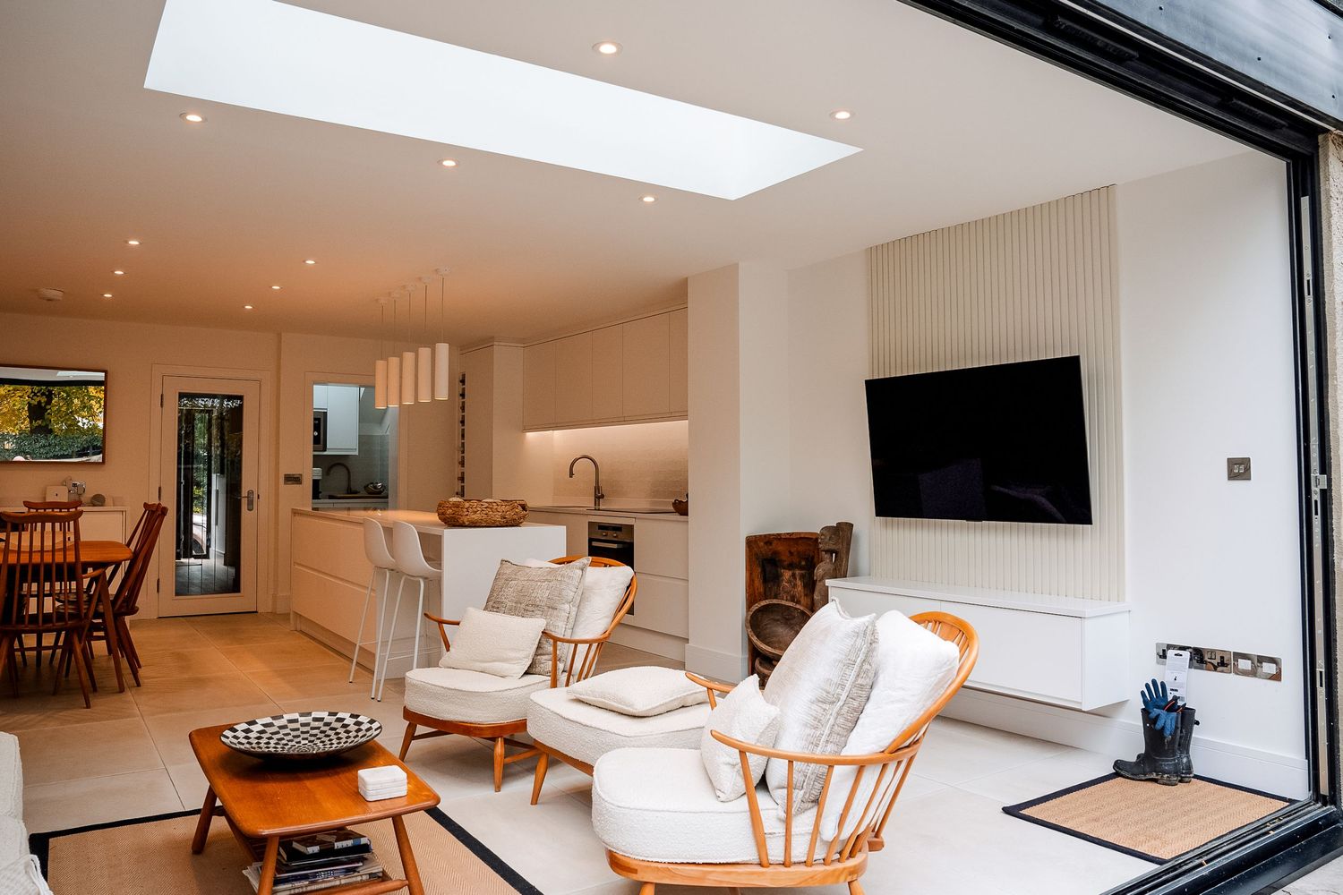 Modern open-plan living room with skylight, white armchairs, wall-mounted TV, and kitchen area with bifold doors.