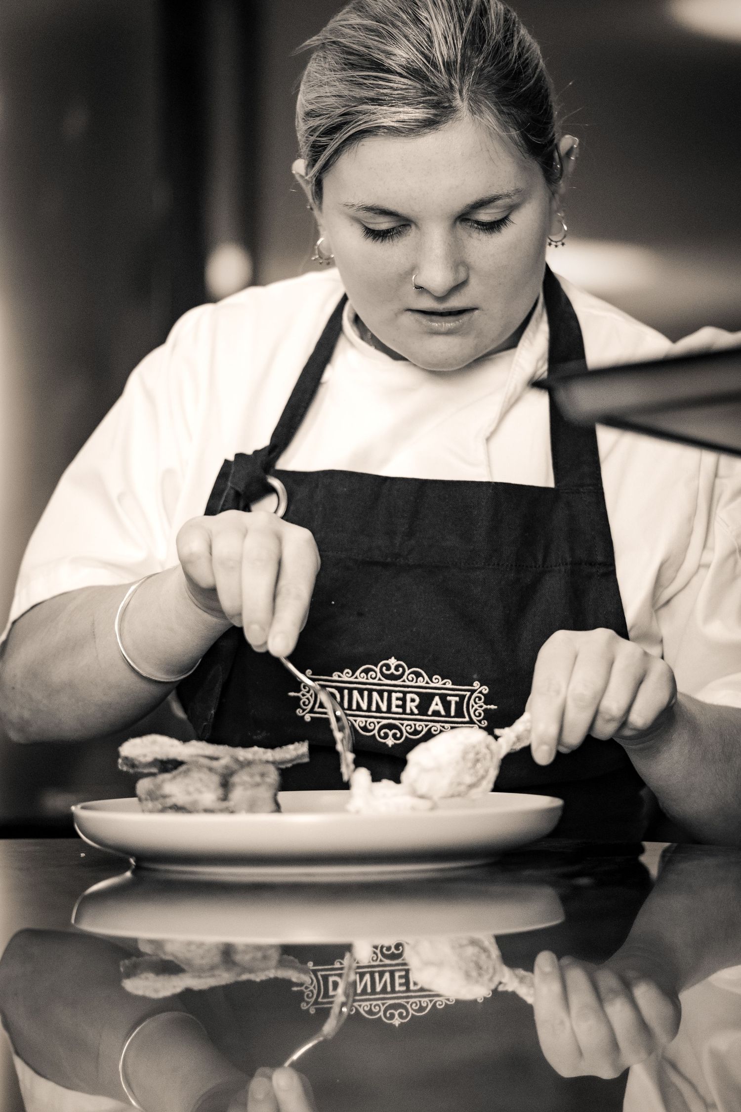 Female chef in black apron carefully plating a dessert dish in a professional kitchen, black and white photo.