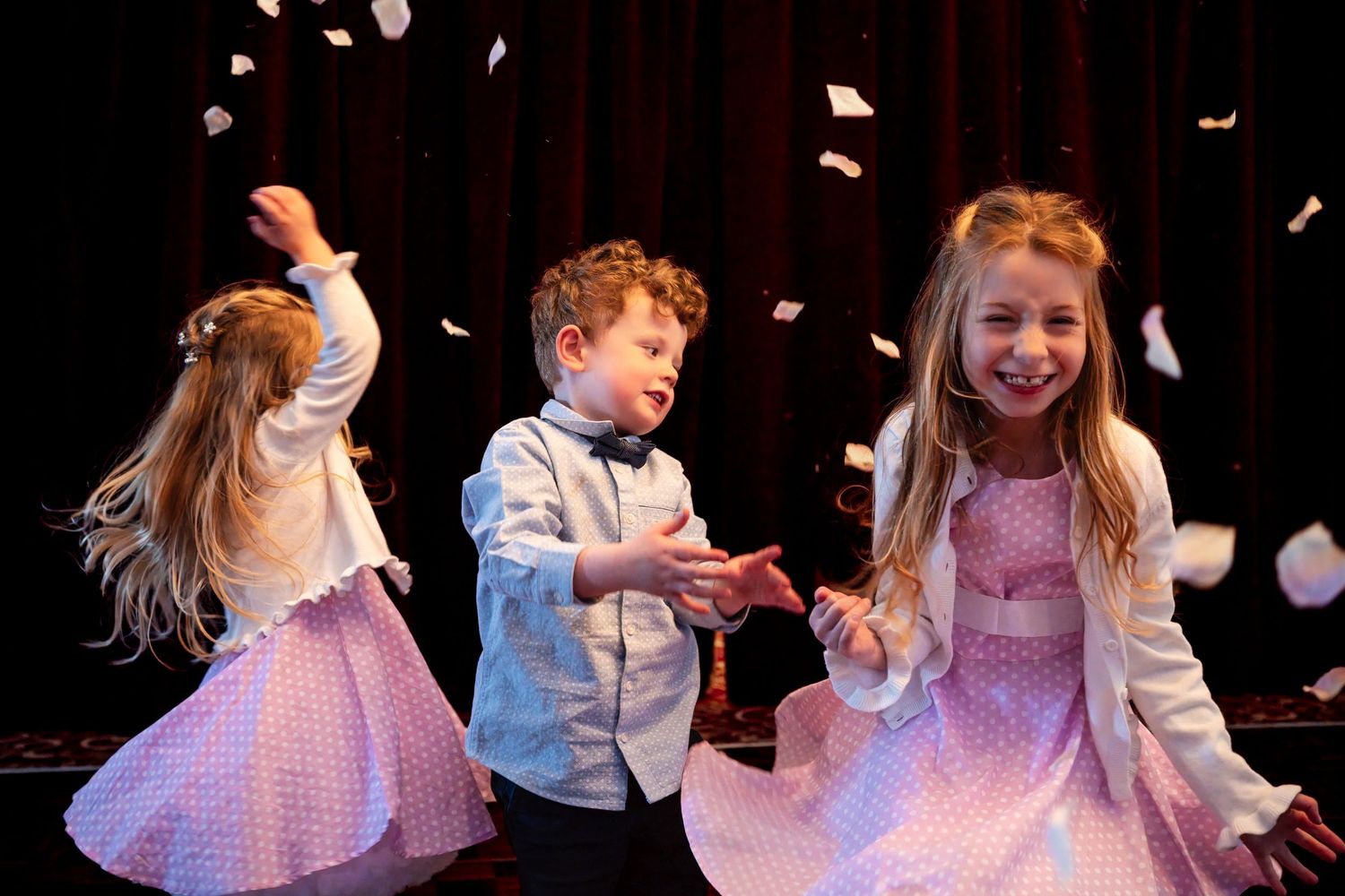 Three children dancing joyfully on stage with confetti falling around them during a performance.