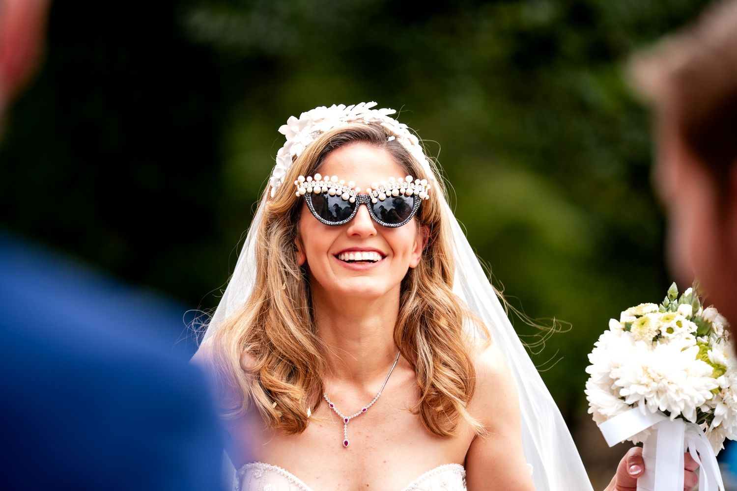 Smiling bride wearing sunglasses and floral crown holds white bouquet outdoors on her wedding day.