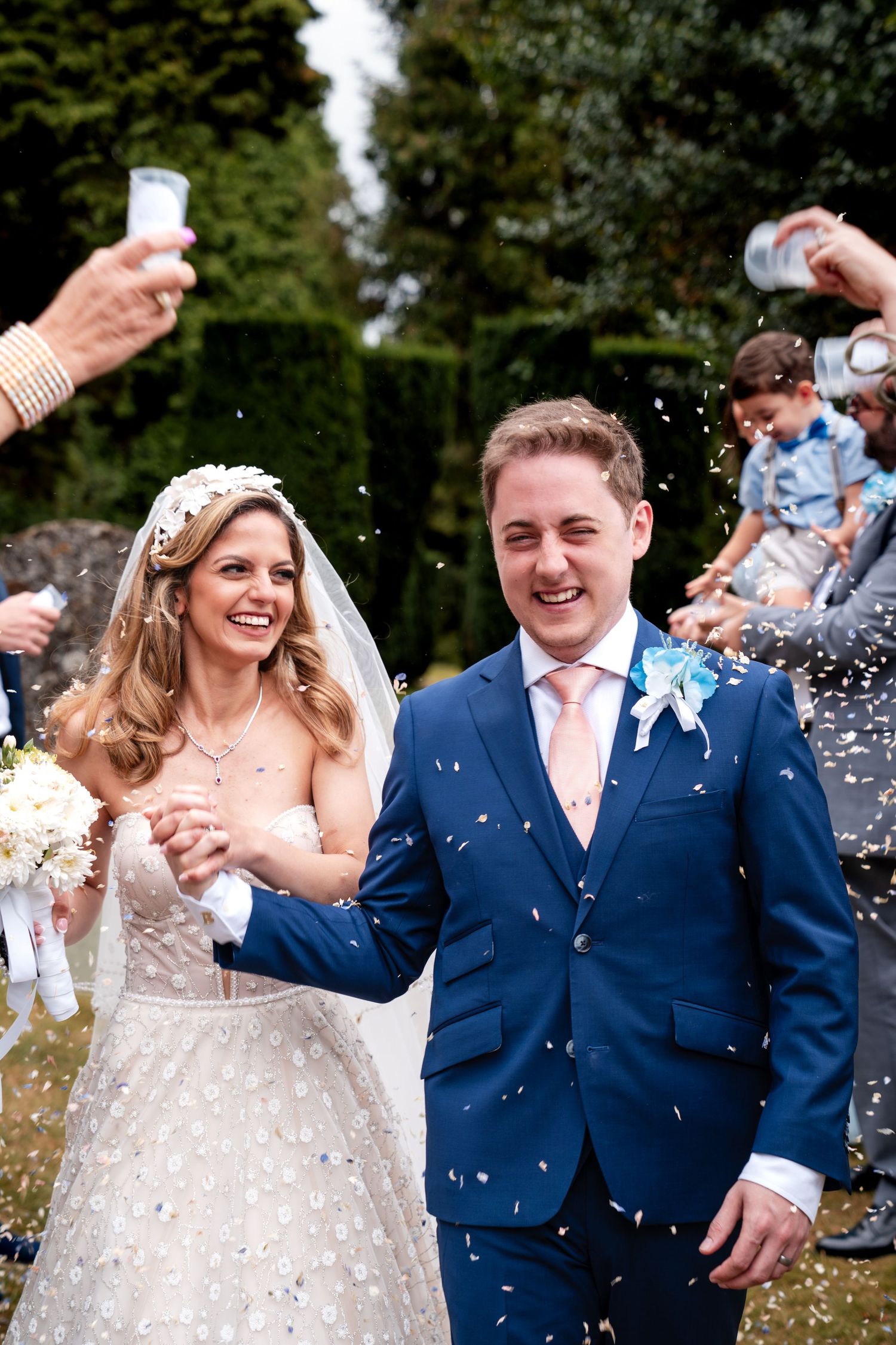 Happy newlyweds walk through confetti shower, bride in white gown with bouquet, groom in blue suit with pink tie.
