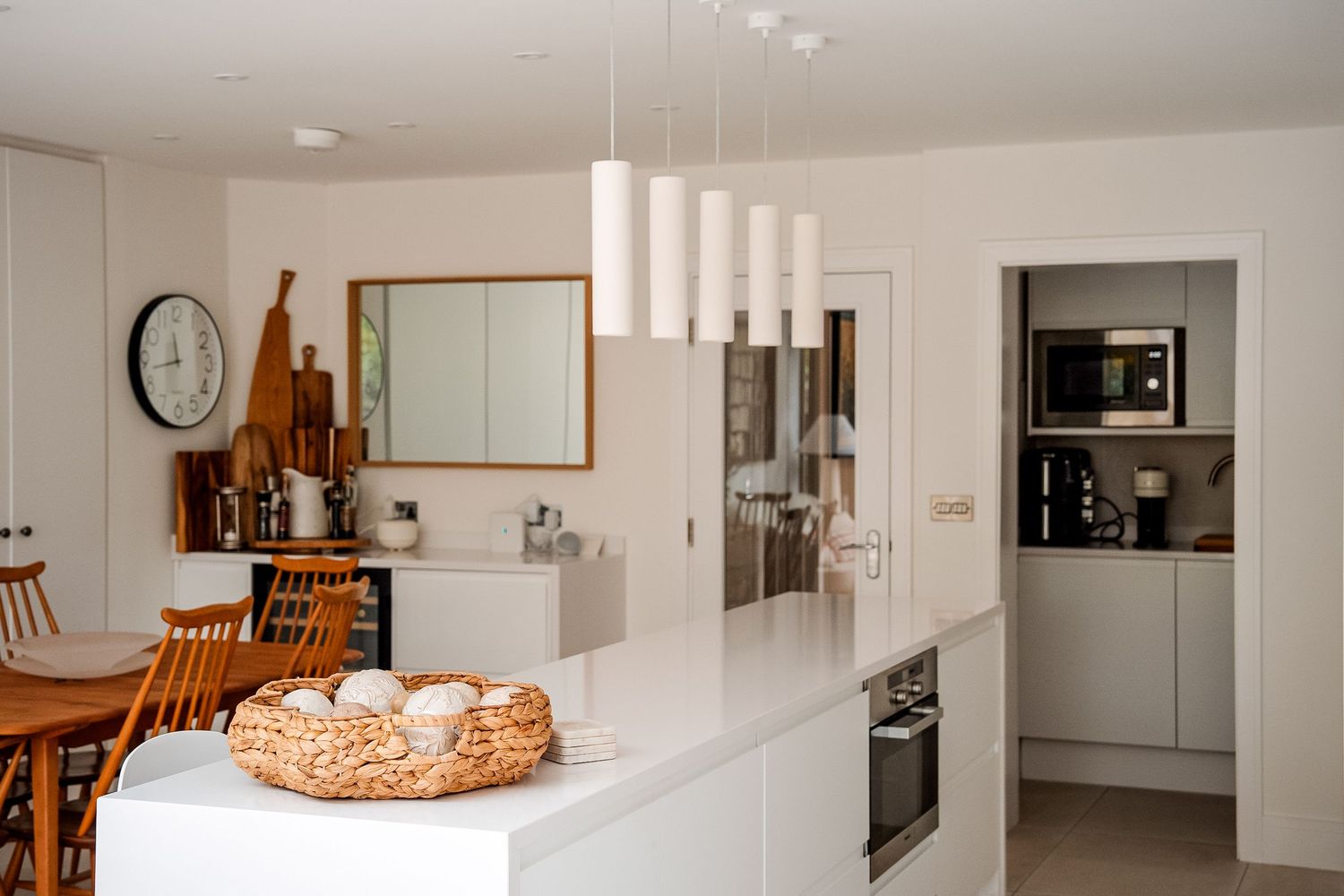 Modern white kitchen with pendant lights, island, dining area, and built-in appliances in a bright open-plan layout.