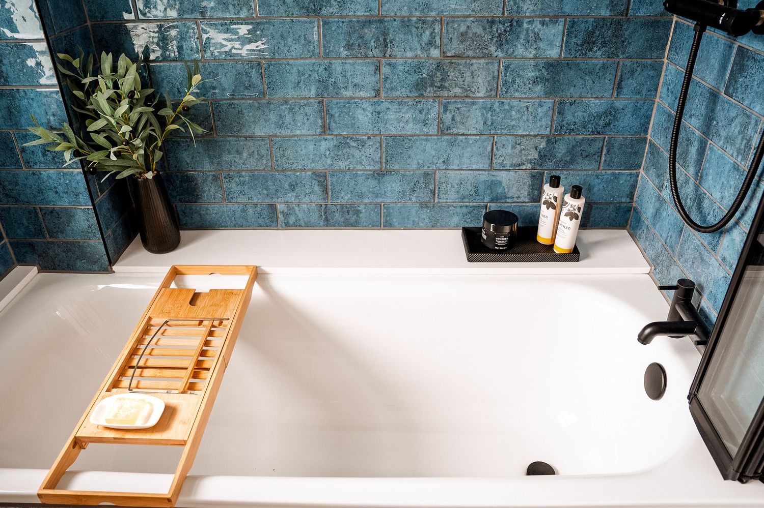 White bathtub with bamboo tray, soap, and toiletries against blue textured tiles with a black shower fixture.
