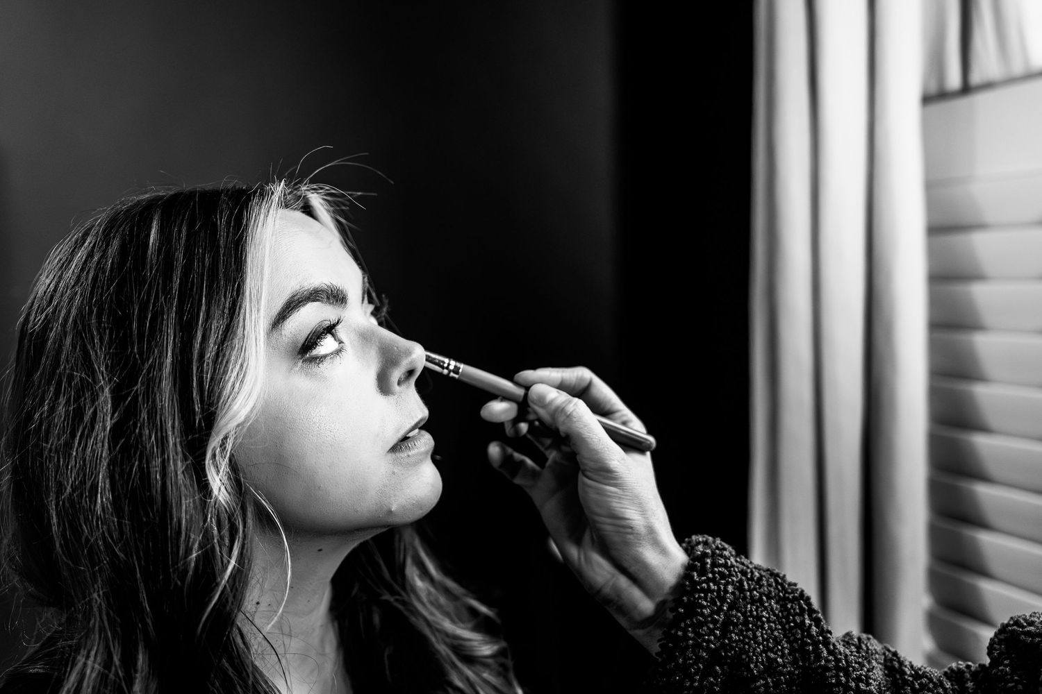 Black and white photo of a woman getting makeup applied with a brush near her nose by a makeup artist.