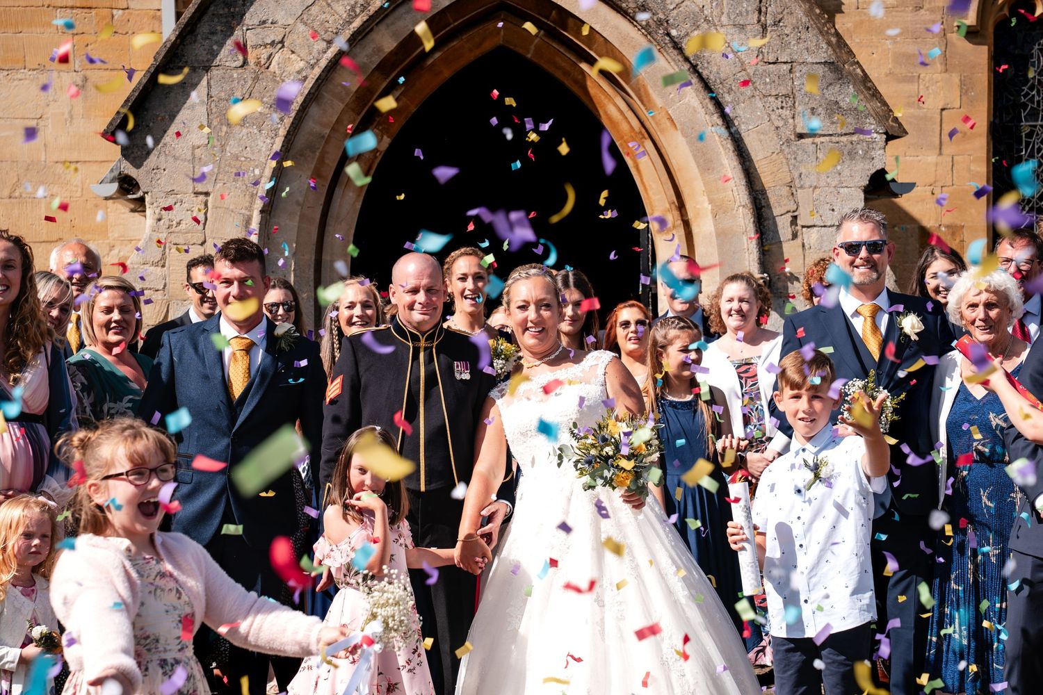 Bride and groom exit stone church amid colorful confetti shower, surrounded by smiling guests and children on sunny wedding day.