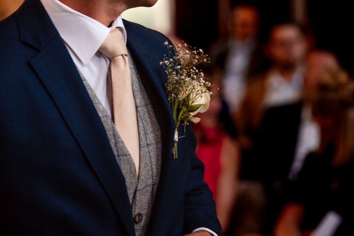 Groom in navy suit with gray vest, blush tie, and delicate white floral boutonniere at a wedding ceremony.
