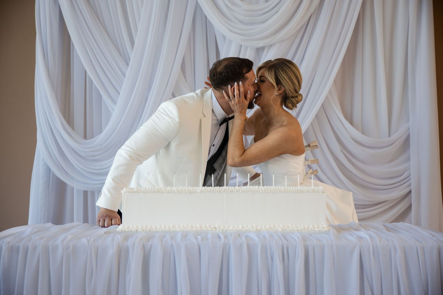 Bride and groom sharing a kiss behind a white wedding cake in front of elegant white draped backdrop.