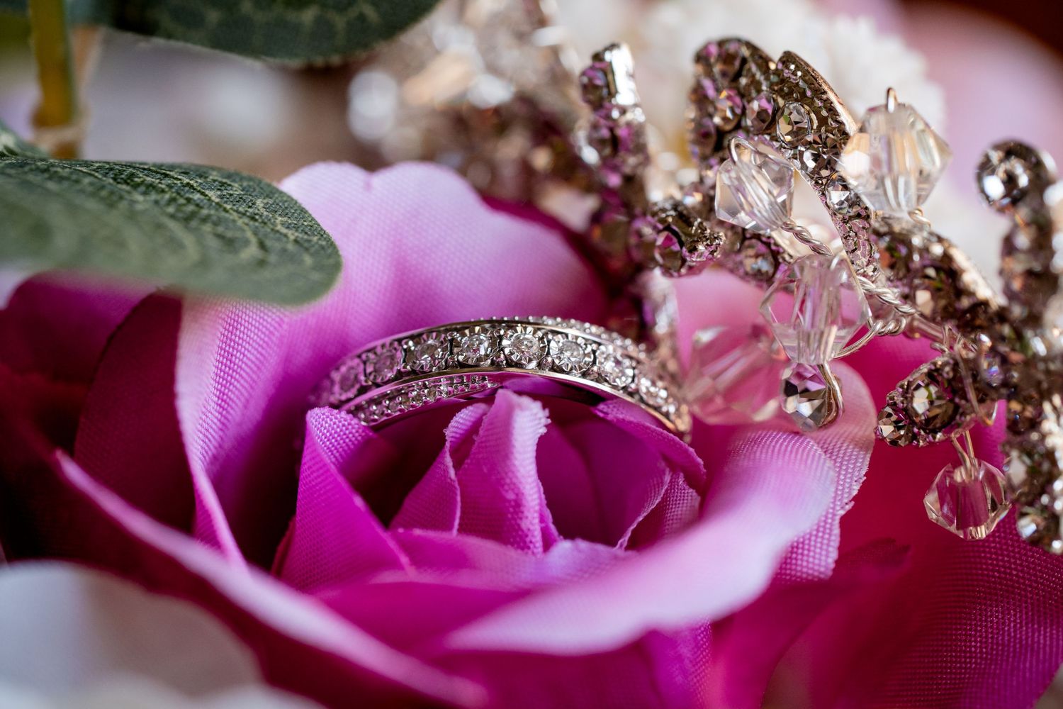 Diamond wedding band ring resting inside a pink rose with crystal jewelry accents in the background.
