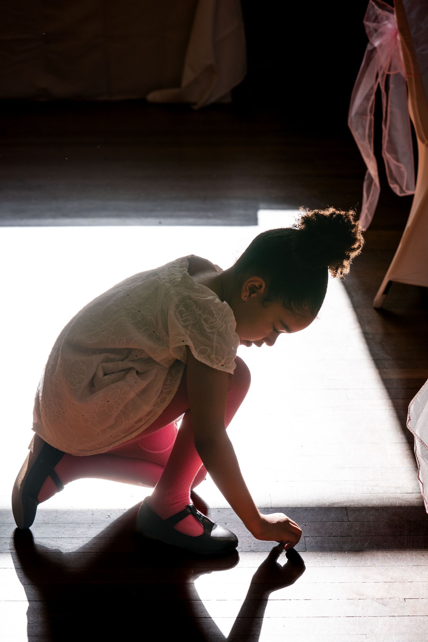 Young girl in white dress and pink tights crouching on wooden floor, putting on ballet shoes in soft backlight.