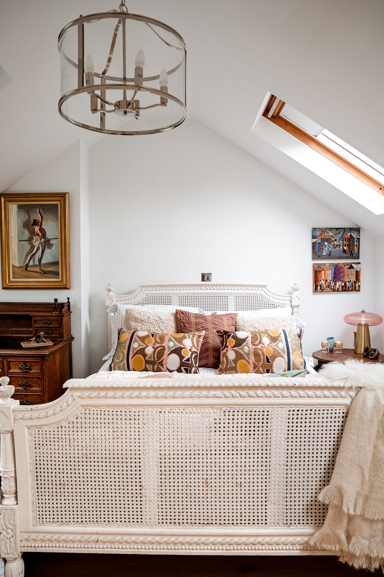 Cozy attic bedroom with white cane bed, eclectic pillows, skylight, chandelier, and vintage artwork on white walls.