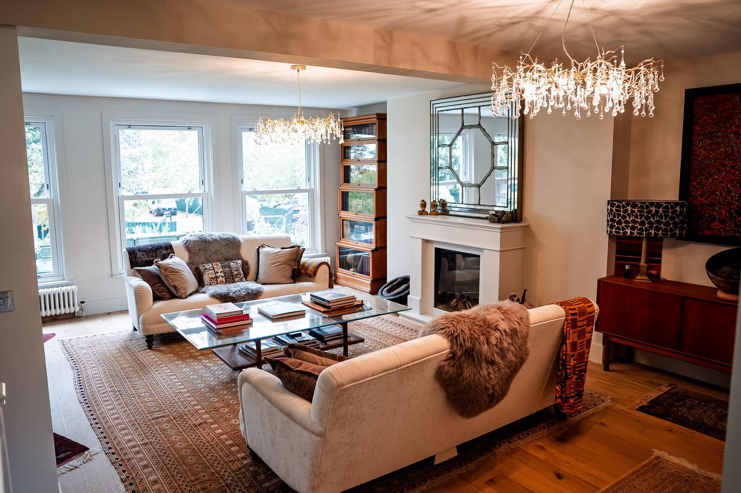 Elegant living room with white sofas, Persian rugs, crystal chandeliers, and a classic fireplace in a bright, stylish interior.