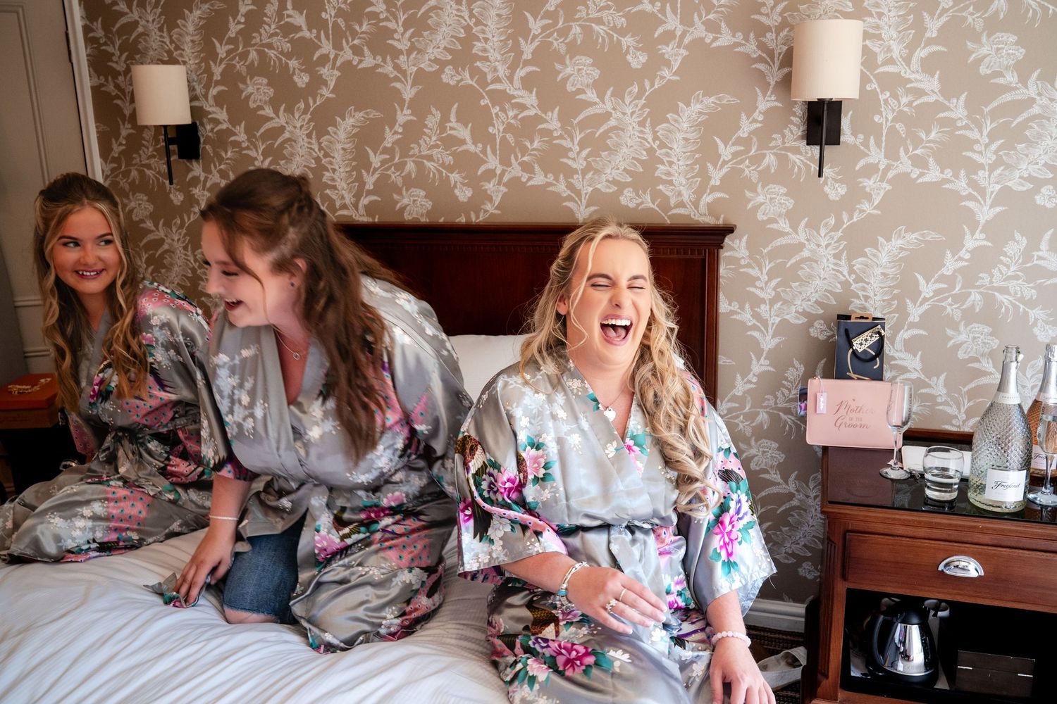 Three bridesmaids laughing joyfully in floral robes while sitting on a hotel bed during wedding preparations.