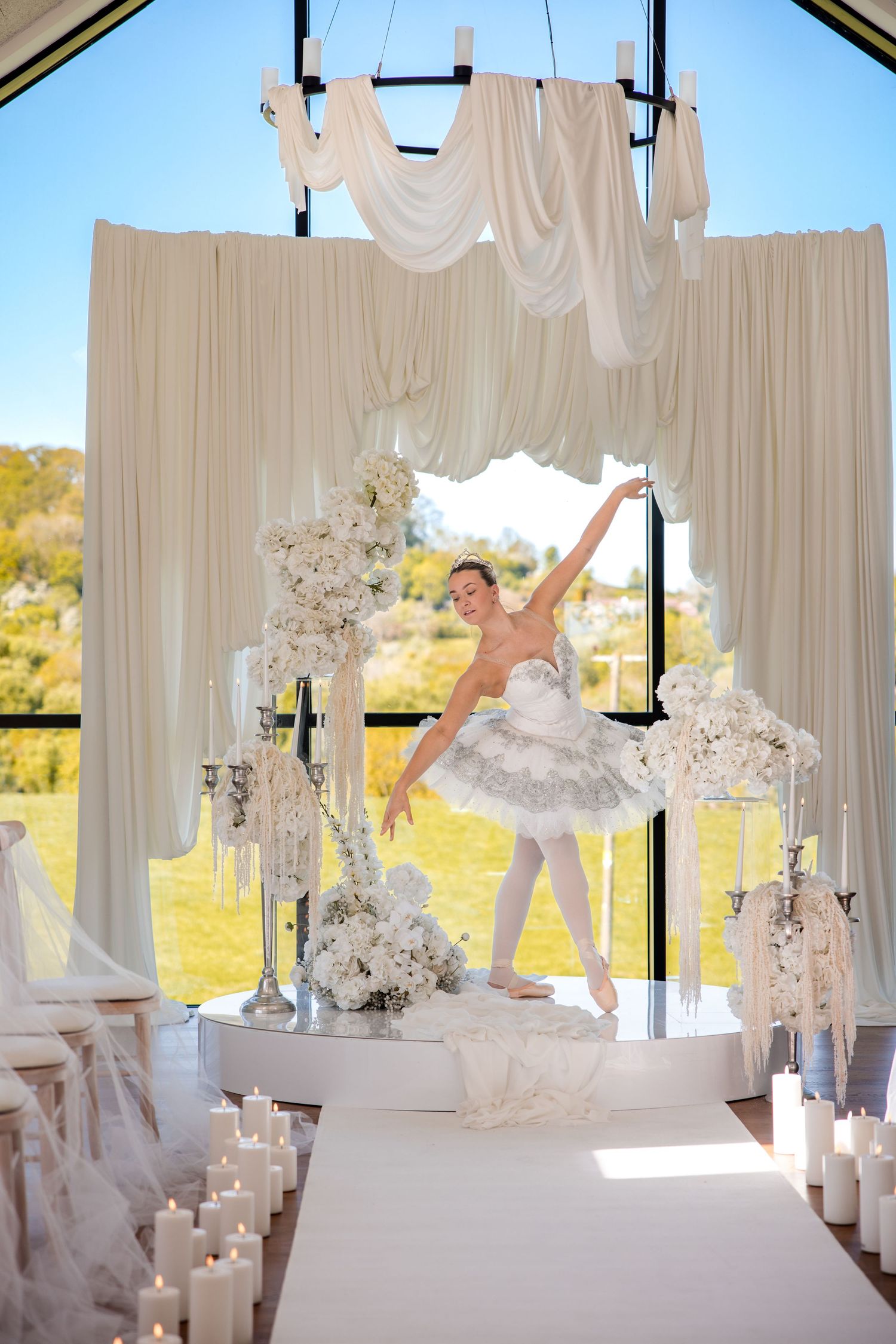 Ballerina in white tutu performing graceful pose on stage adorned with candles and floral arrangements at elegant wedding venue.
