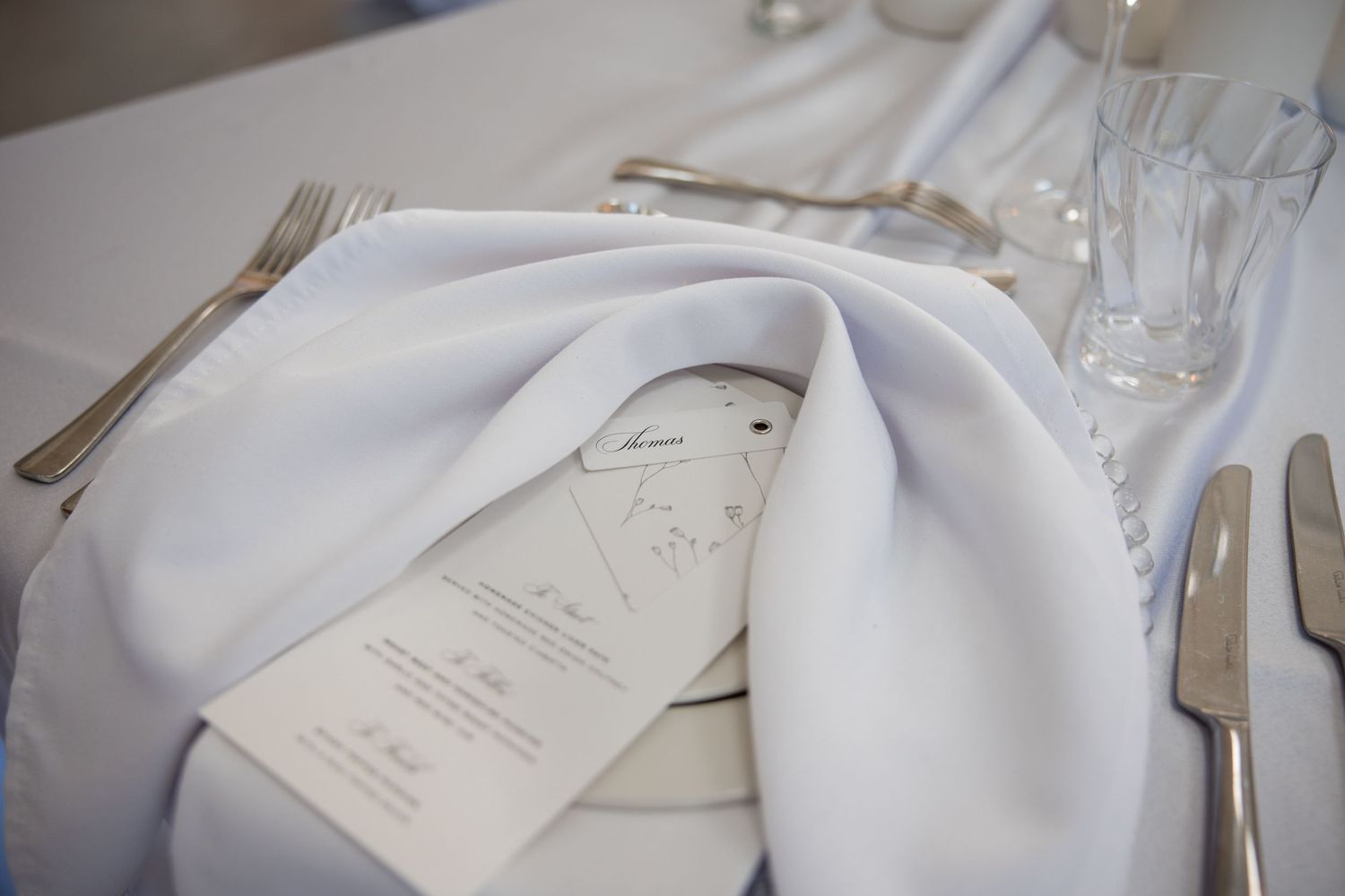 Elegant white wedding table setting with folded napkin, silverware, crystal glasses, and a decorative menu card.