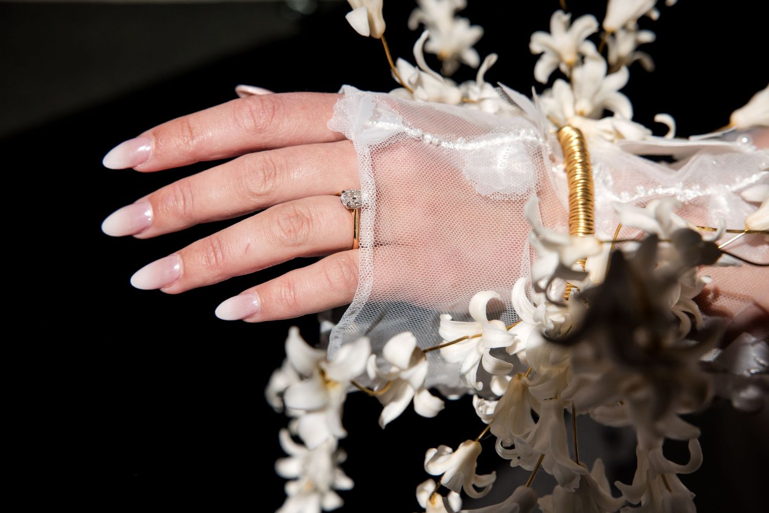 Close-up of a hand with long almond nails wearing a diamond ring, gold bracelet, and white orchids on dark background.