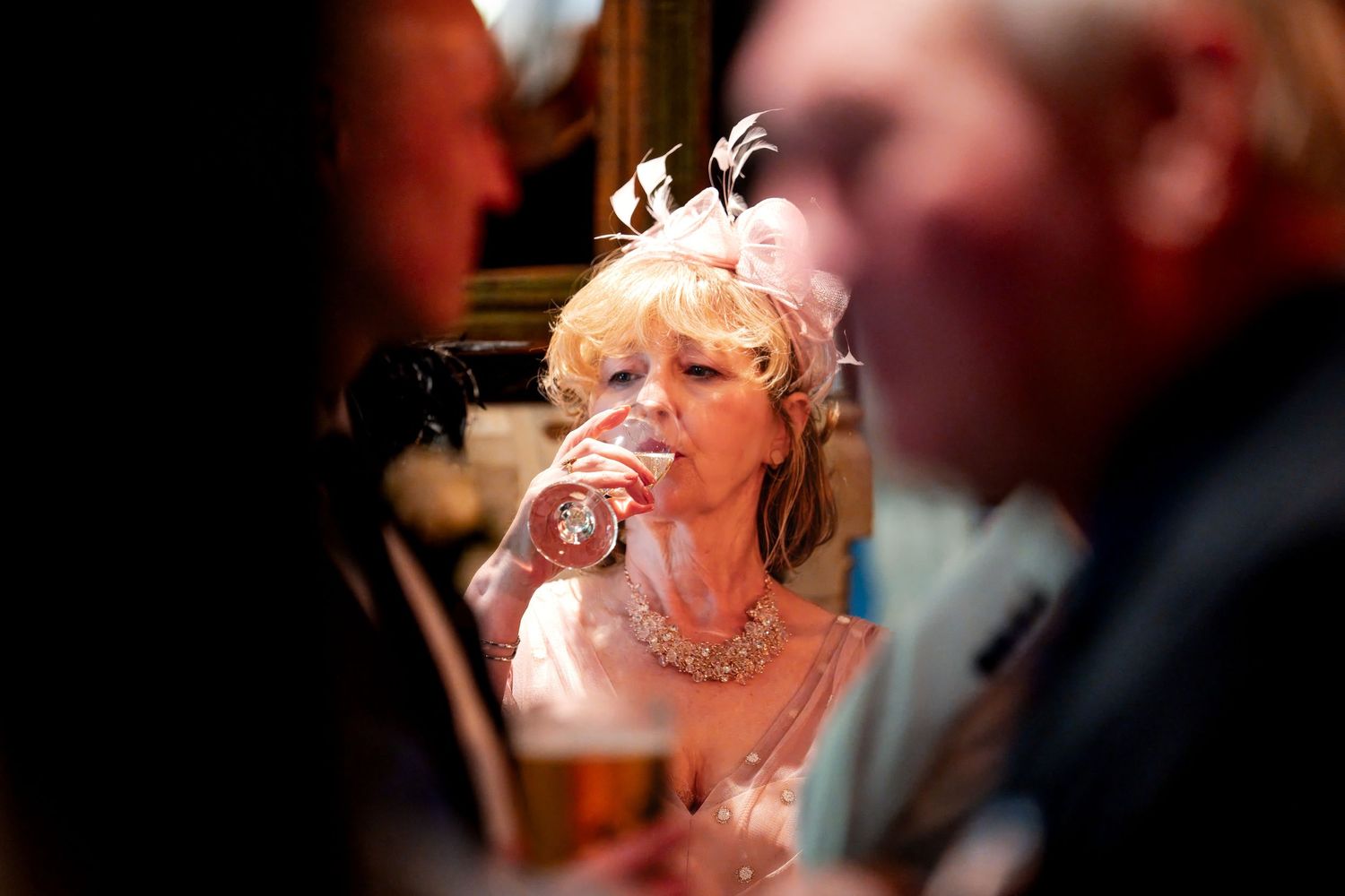 Elegantly dressed woman in pink with feathered fascinator drinking from a glass at a formal social event.