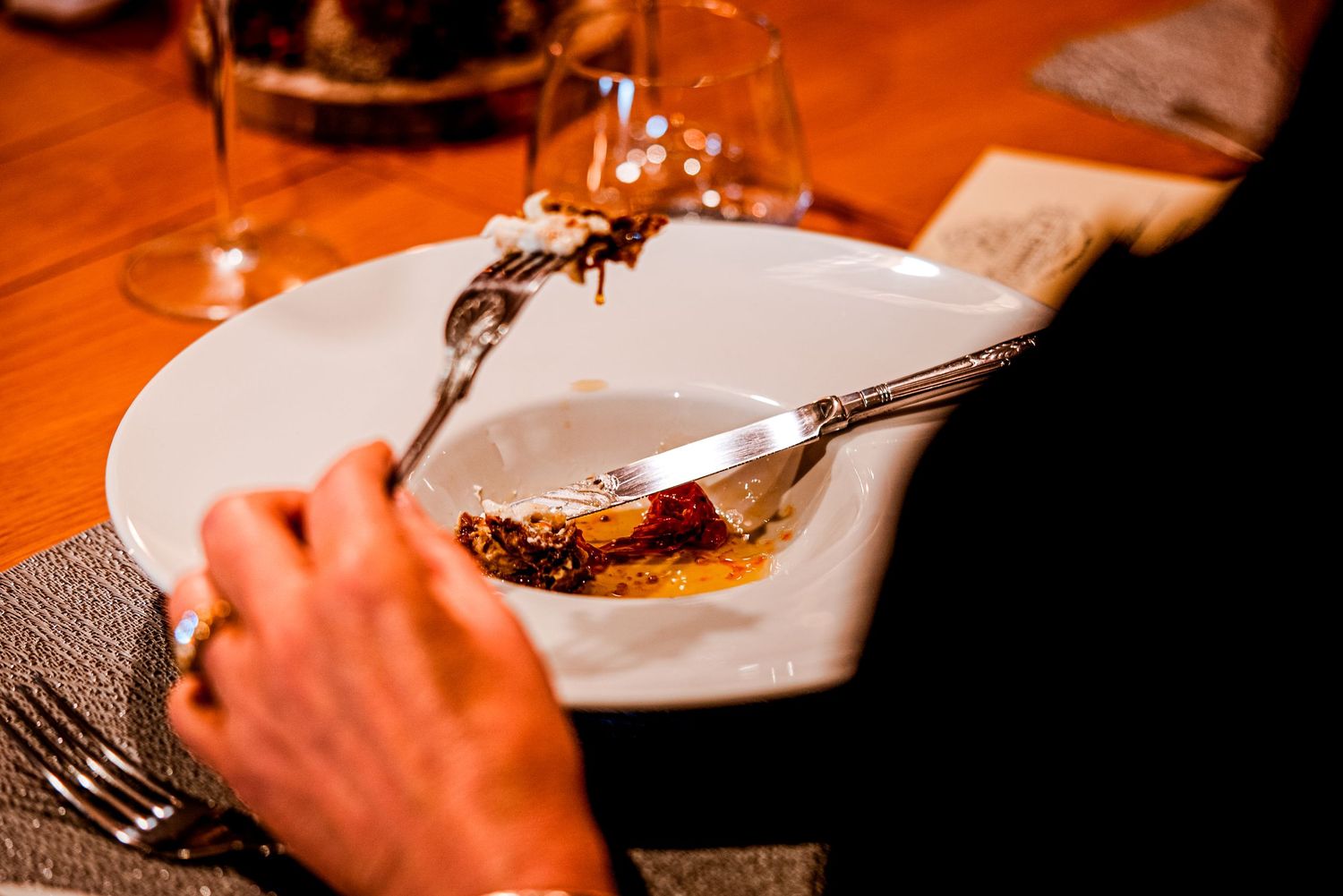 Person cutting into a saucy meat dish on a white plate at a wooden restaurant table with wine glasses.