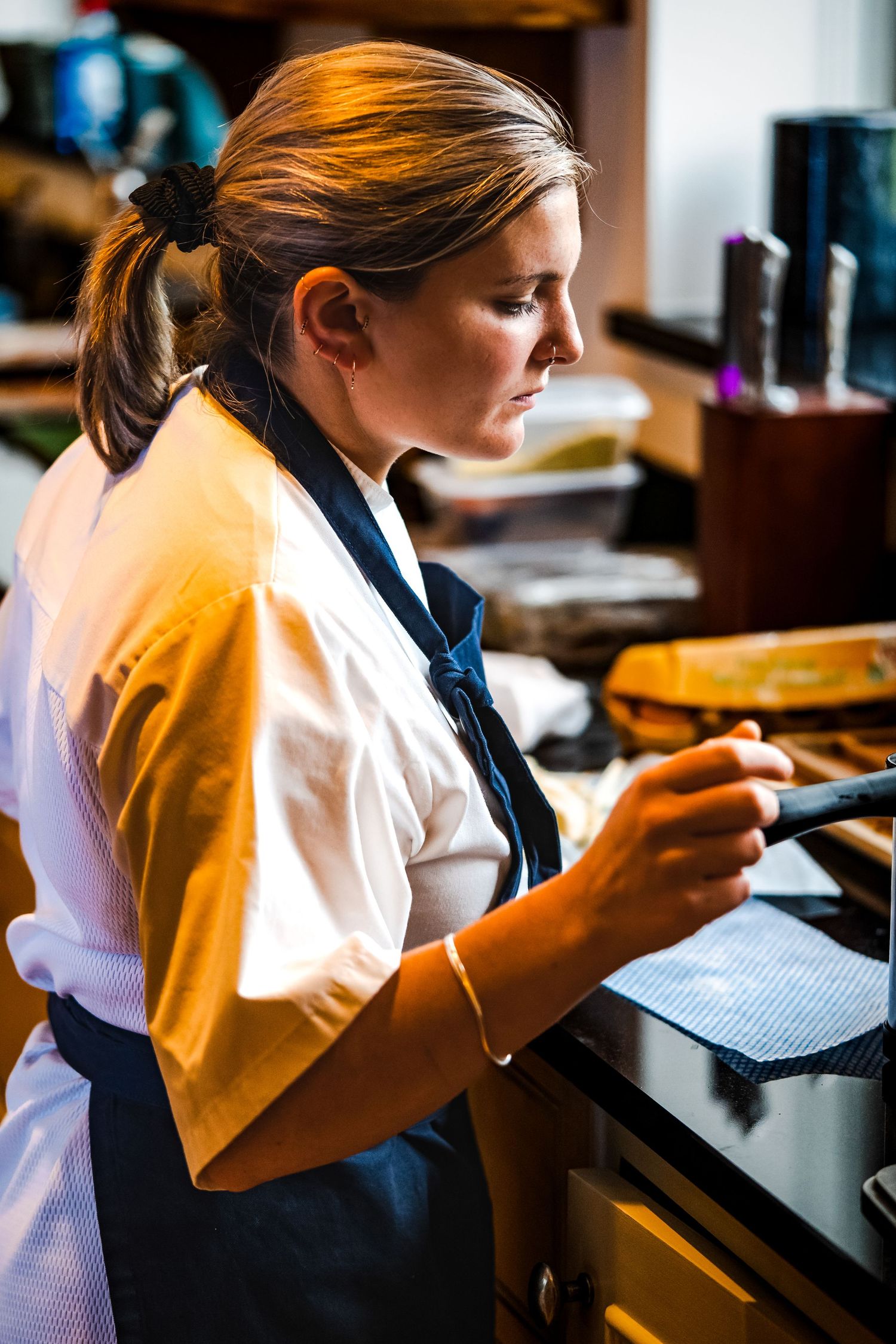 Female chef in white shirt and dark apron carefully examining food preparation in a professional kitchen.