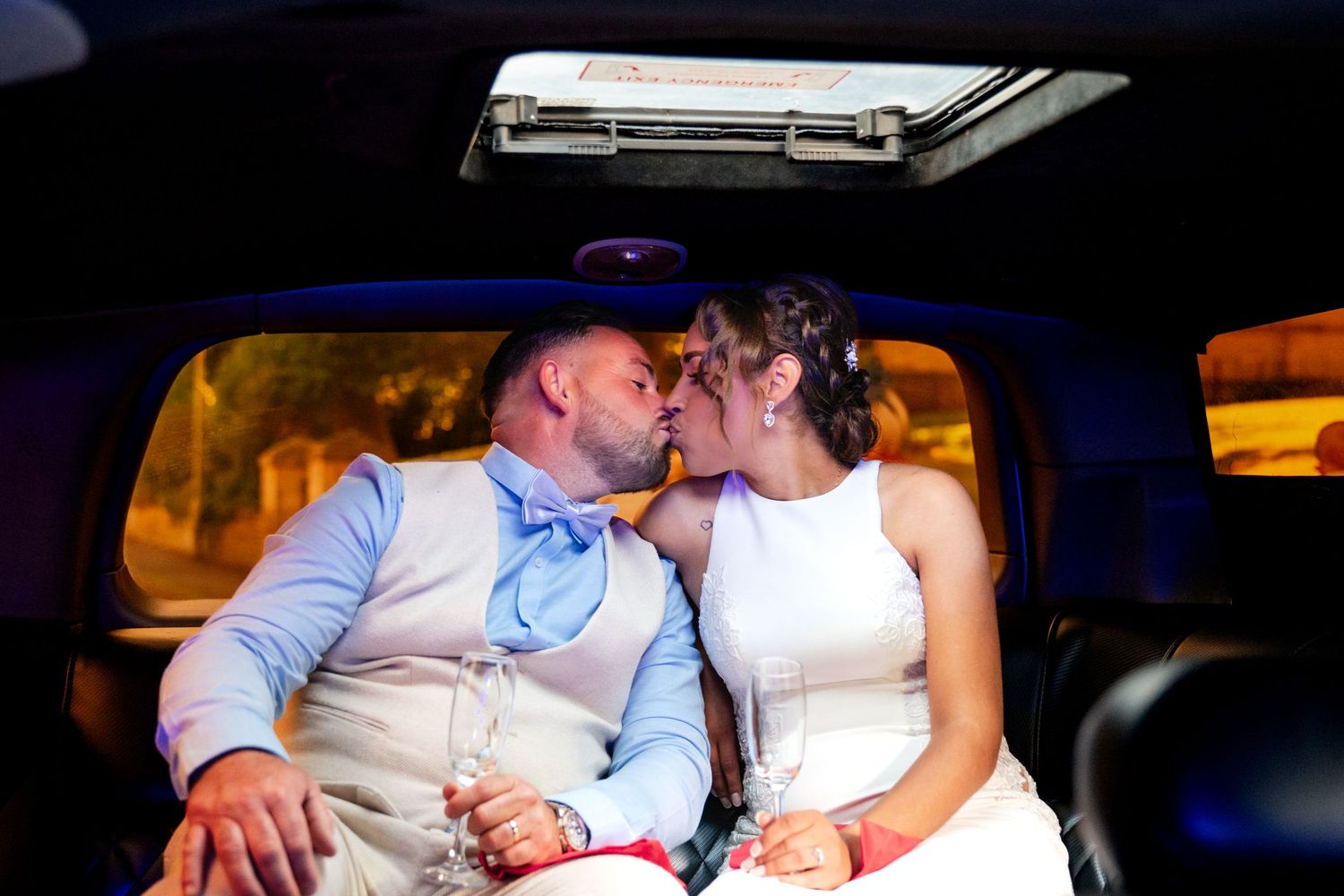 Bride and groom sharing a romantic kiss inside a limousine, dressed in wedding attire with champagne glasses.