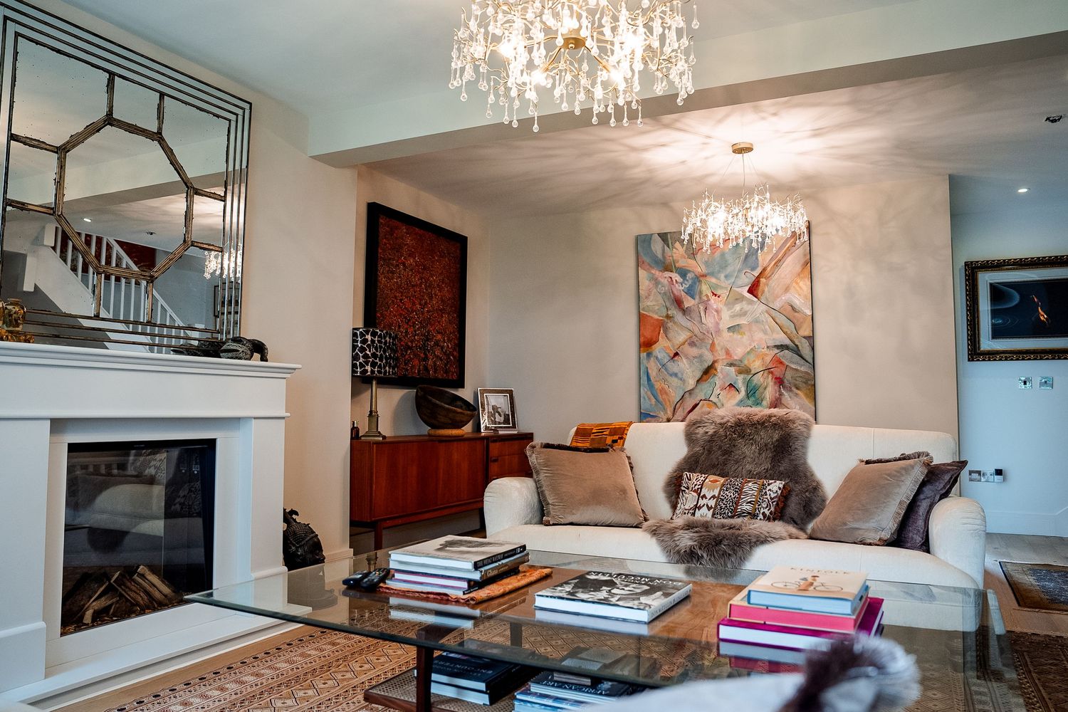 Elegant living room with crystal chandelier, white sofas, glass coffee table, fireplace, and colorful abstract artwork on walls.