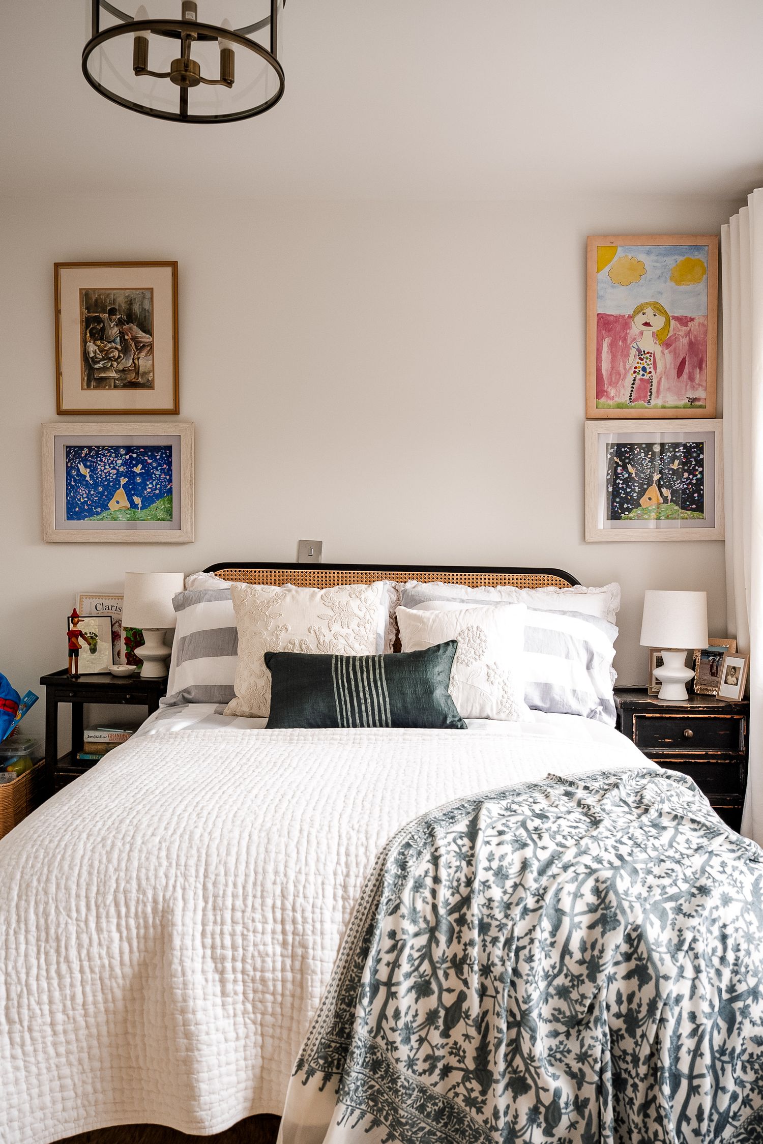 Cozy bedroom with white quilt, green throw, rattan headboard, black nightstands, and colorful children's artwork on walls.