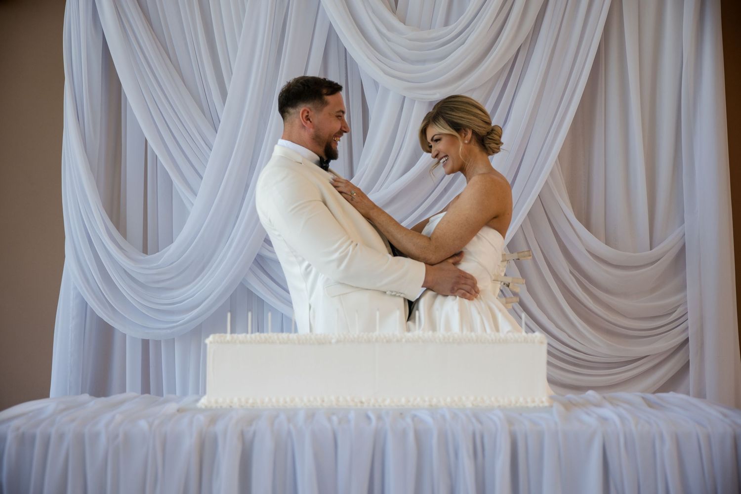 Bride and groom embrace behind white wedding cake with elegant draped white fabric backdrop.