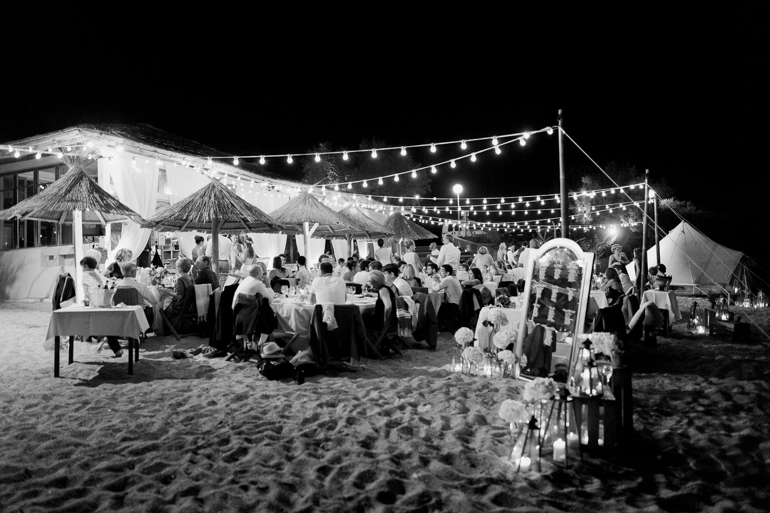 Photo en noir et blanc d'une élégante réception de mariage sur la plage, de nuit, avec des guirlandes lumineuses  à calvi pain de sucre