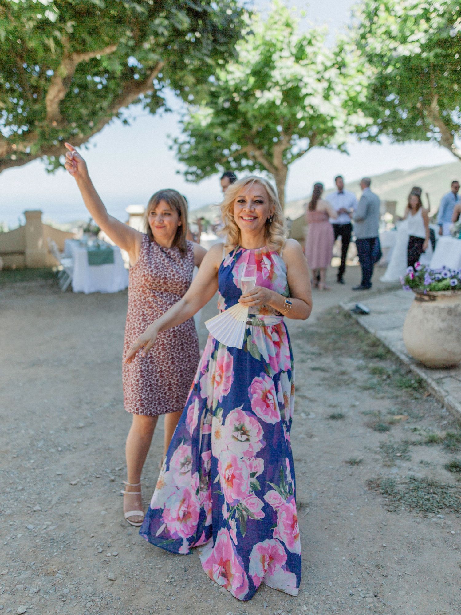 Two women in floral dresses smile at an outdoor wedding reception under shady trees on a sunny day.