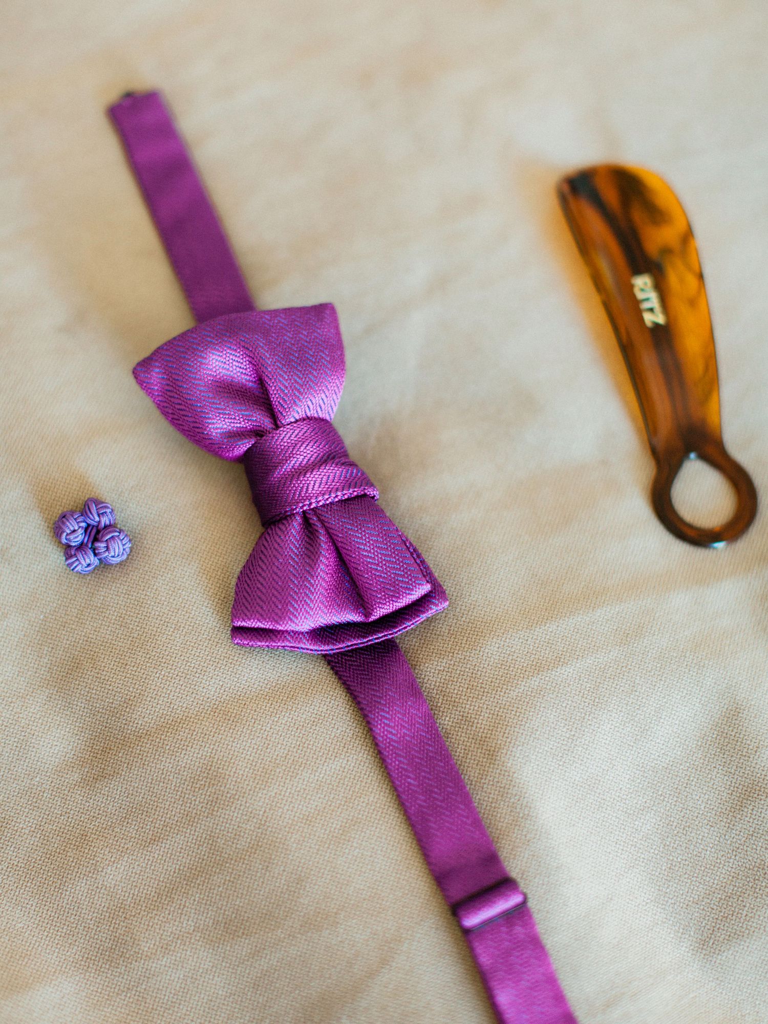 Purple satin bow tie laid flat with matching cufflinks and a tortoiseshell hair clip on a neutral background.