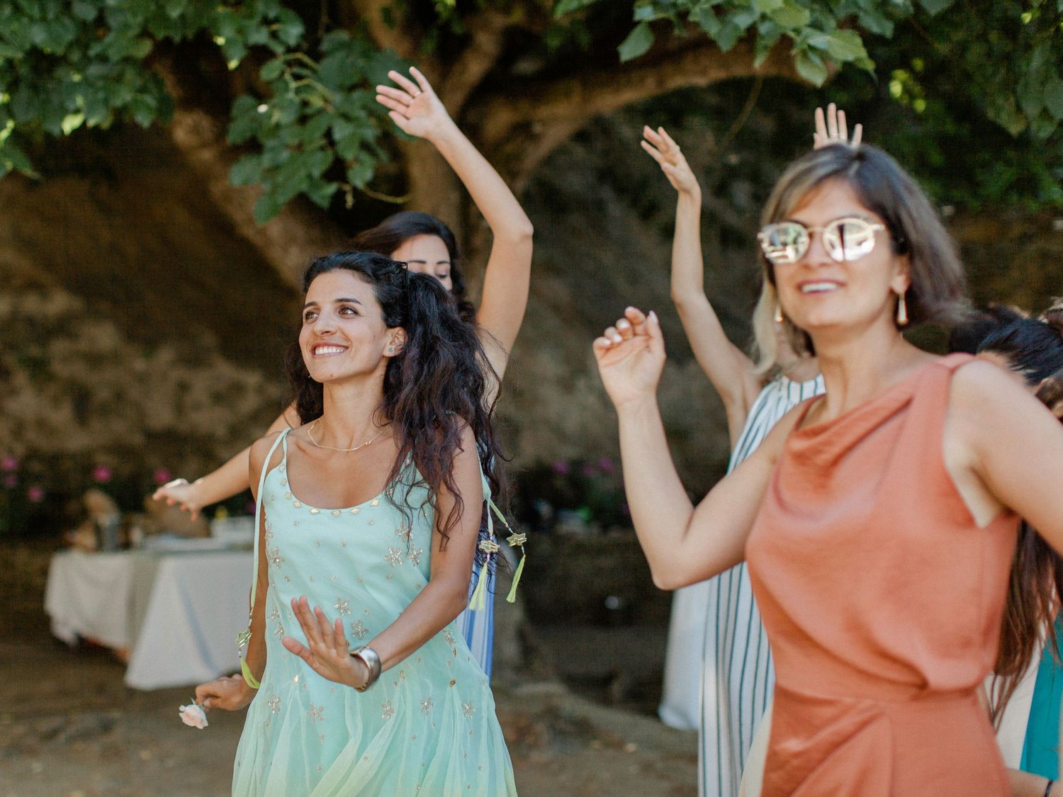 Women dancing outdoors at a garden party near a rustic tree, wearing colorful summer dresses.