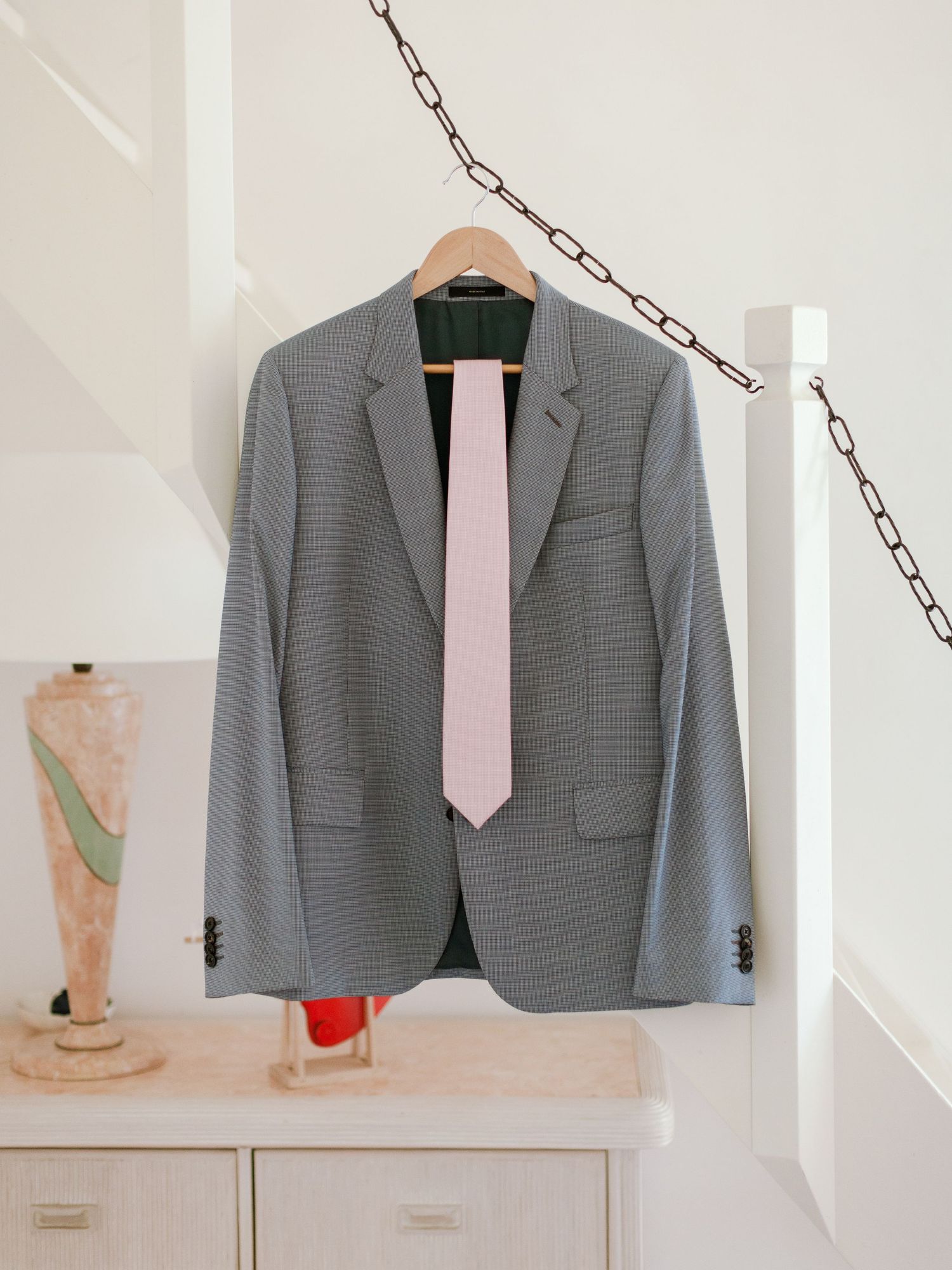Blazer gris à motif pied-de-poule avec une cravate rose sur un cintre en bois, présenté sur un mur blanc au-dessus d'une commode.