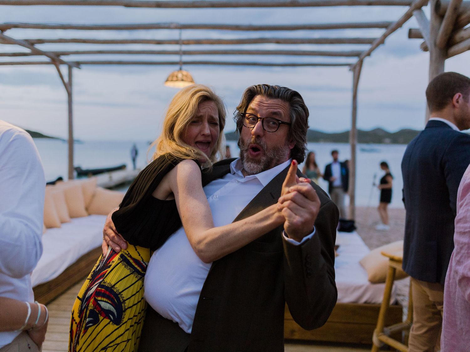 Un couple danse enlacé lors d'un événement en plein air sur la plage au crépuscule, la femme en robe colorée, l'homme en blazer.