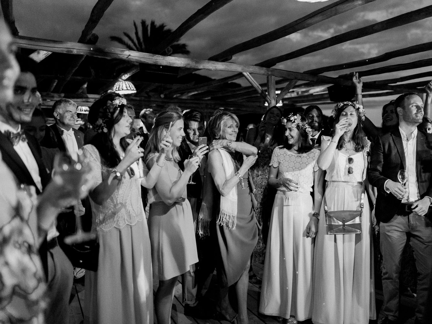 Photo en noir et blanc d'invités élégamment vêtus, dansant et discutant lors d'une réception de mariage en plein air.